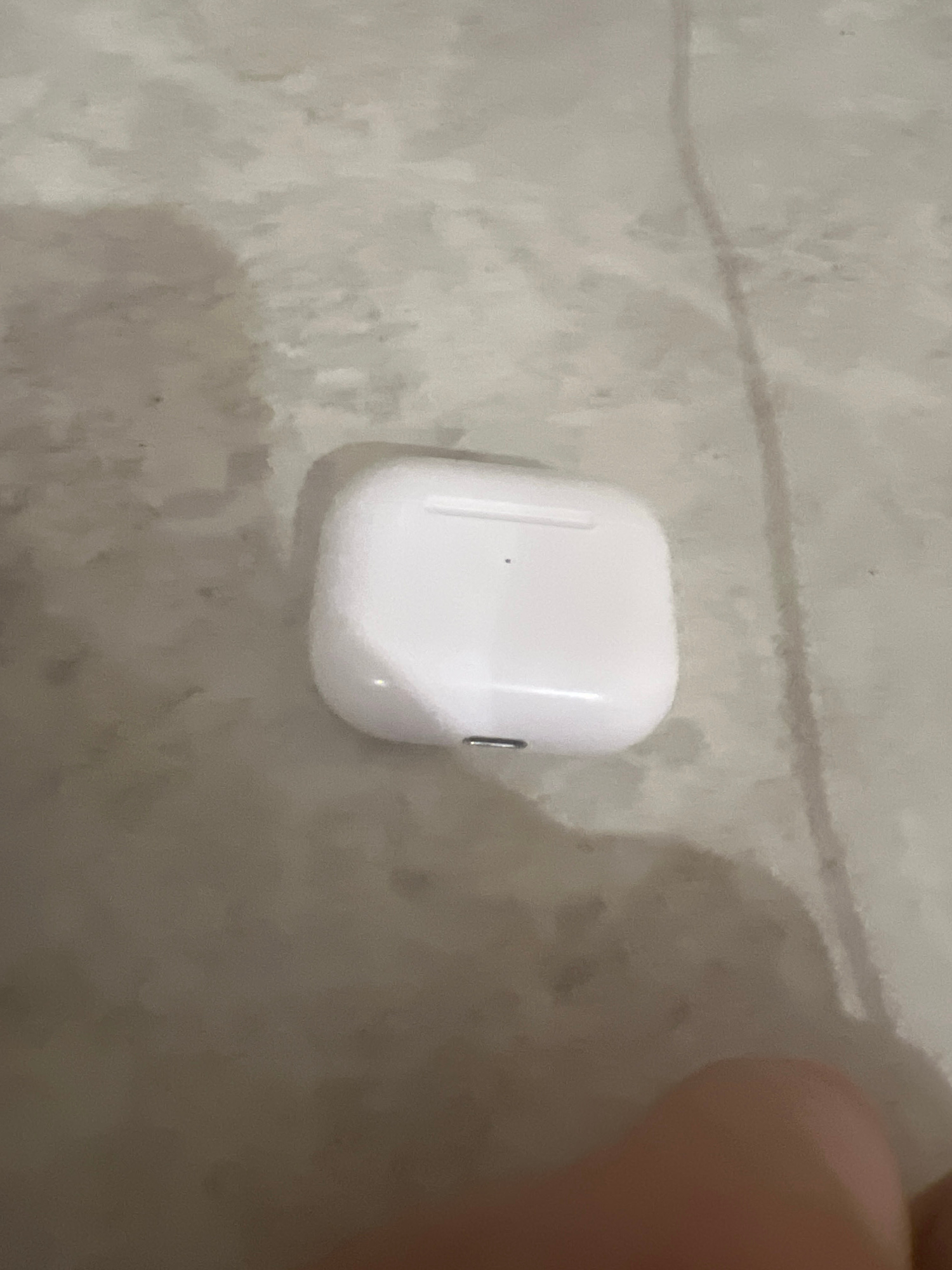 A white, rectangular charging case, likely for wireless earbuds, rests on a light-colored, textured surface. A small, dark circular dot is visible on the top surface of the case, and a silver charging port is located on the bottom front. The surface appears to be a floor or countertop, with subtle variations in color and texture, possibly indicating a lightly mottled or stained material. Shadows are cast on the surface, suggesting an overhead light source. A portion of a person's hand or finger, with visible skin tone, is visible in the lower right corner of the frame, partially obscuring the background. The overall lighting is dim, contributing to the grainy appearance of the image. This image was taken in Saratov, Russia.