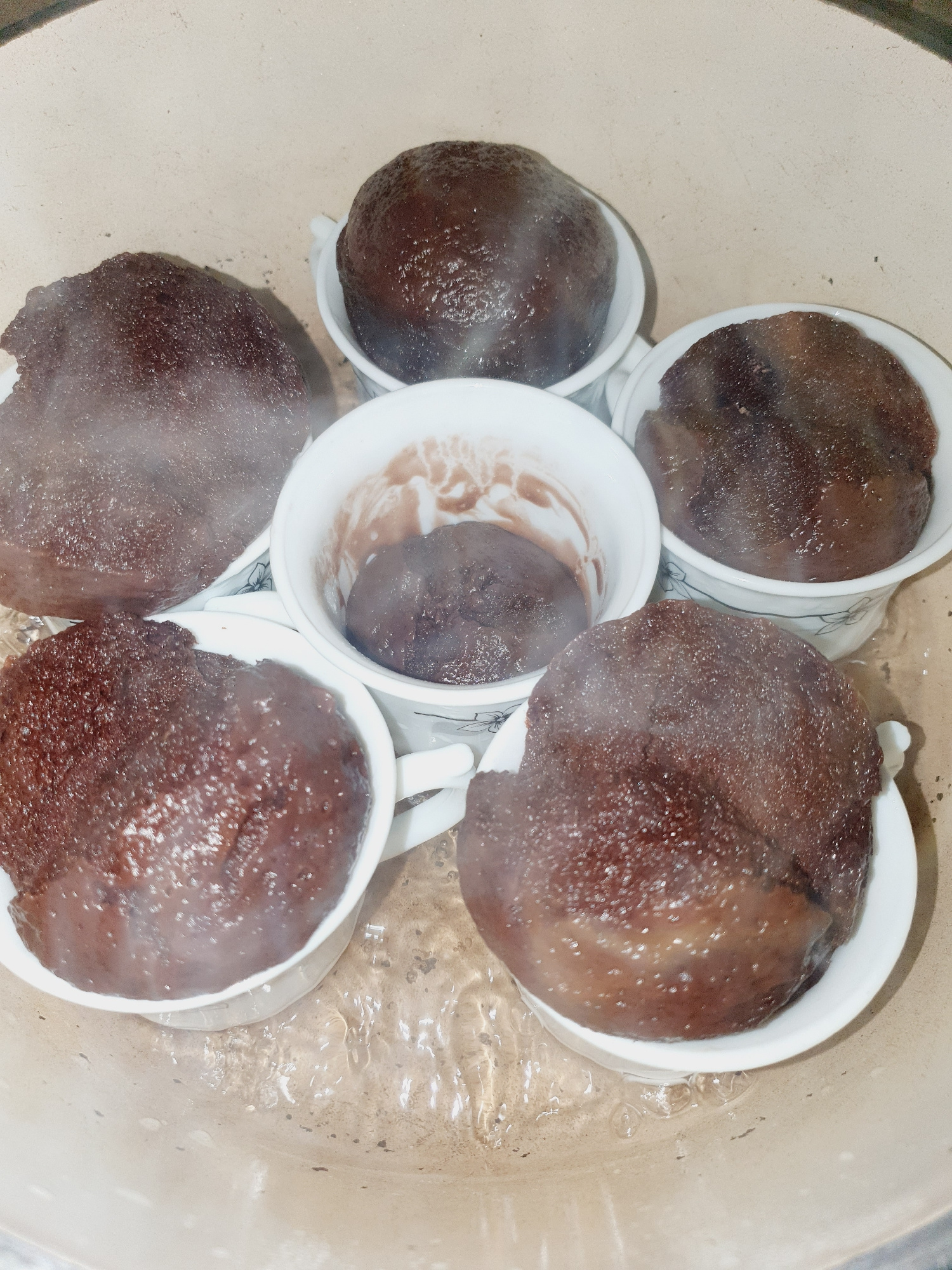 The image shows six small white ceramic cups arranged in a circular pattern within a light-colored pot. Each cup contains a dark brown, spherical food item that appears to be steaming. The food items are partially submerged in a liquid, likely water, which is gently bubbling in the bottom of the pot. The cups have a delicate floral pattern on the inside near the rim. The lighting is dim, and steam is visible rising from some of the food items. There are no people visible in the image. The scene is a close-up, overhead view of the food preparation process. This image does not contain any visual information that would allow for its specific location to be determined within the city of Shiraz, Iran.