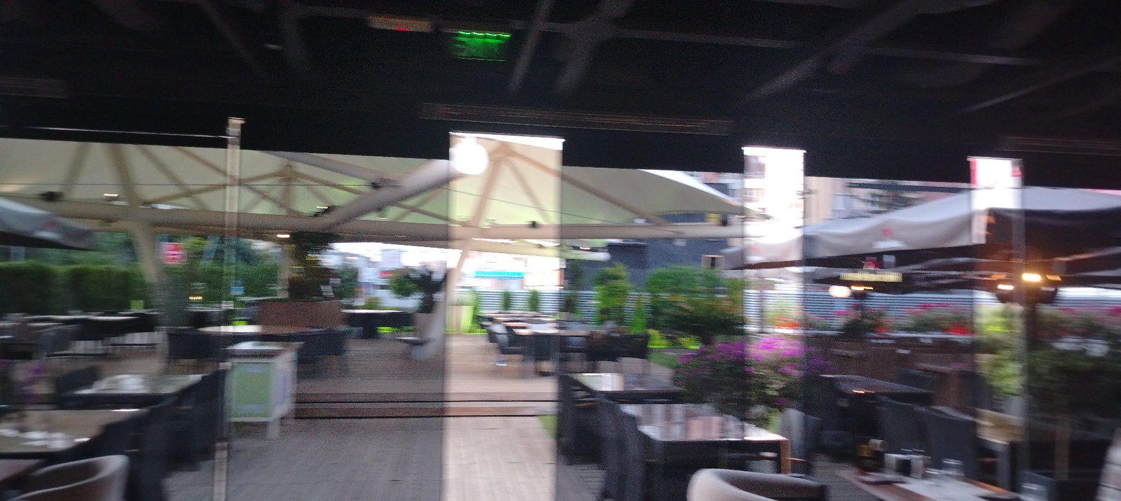 This image shows an outdoor seating area of a restaurant or cafe, likely in Sofia, Bulgaria, with a glass partition in the foreground. The setting appears to be an urban environment, with buildings visible in the background through the glass. The outdoor area features multiple tables and chairs, some set up for dining, with umbrellas or canopies providing shade. There are also some plants and decorative elements, suggesting a pleasant ambiance. The overall impression is of a commercial establishment ready for customers, with the glass partition offering a glimpse into the interior or a view of the exterior. The image quality is slightly blurry, but the general layout and components of the scene are discernible.