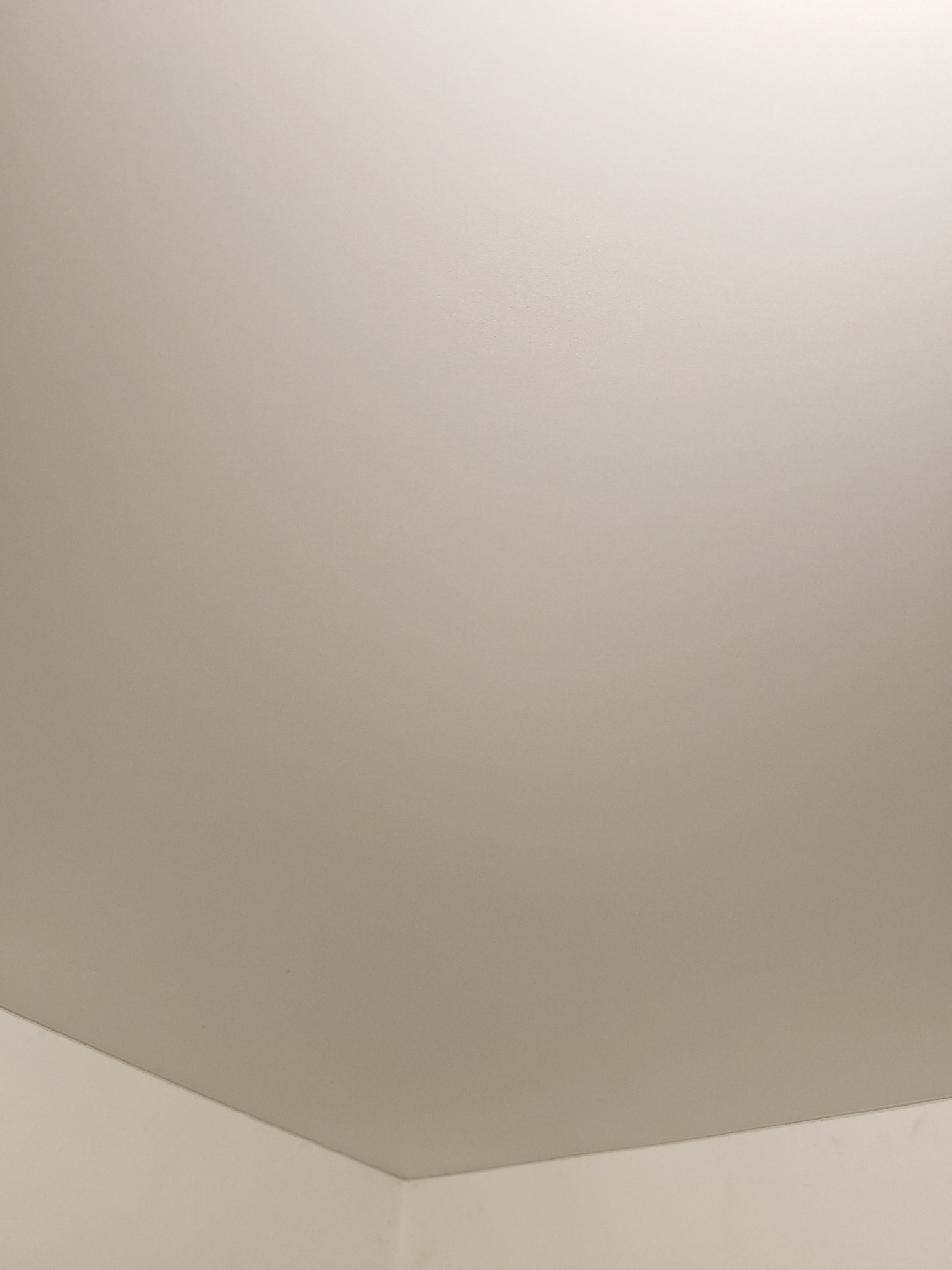 A flat, beige ceiling fills the frame, with a slight gradient indicating light source. In the lower left and lower right corners, a white wall meets the ceiling at a sharp angle. The surfaces appear smooth and unremarkable. There are no people, objects, or distinguishing features visible in the image. The setting is indoors.