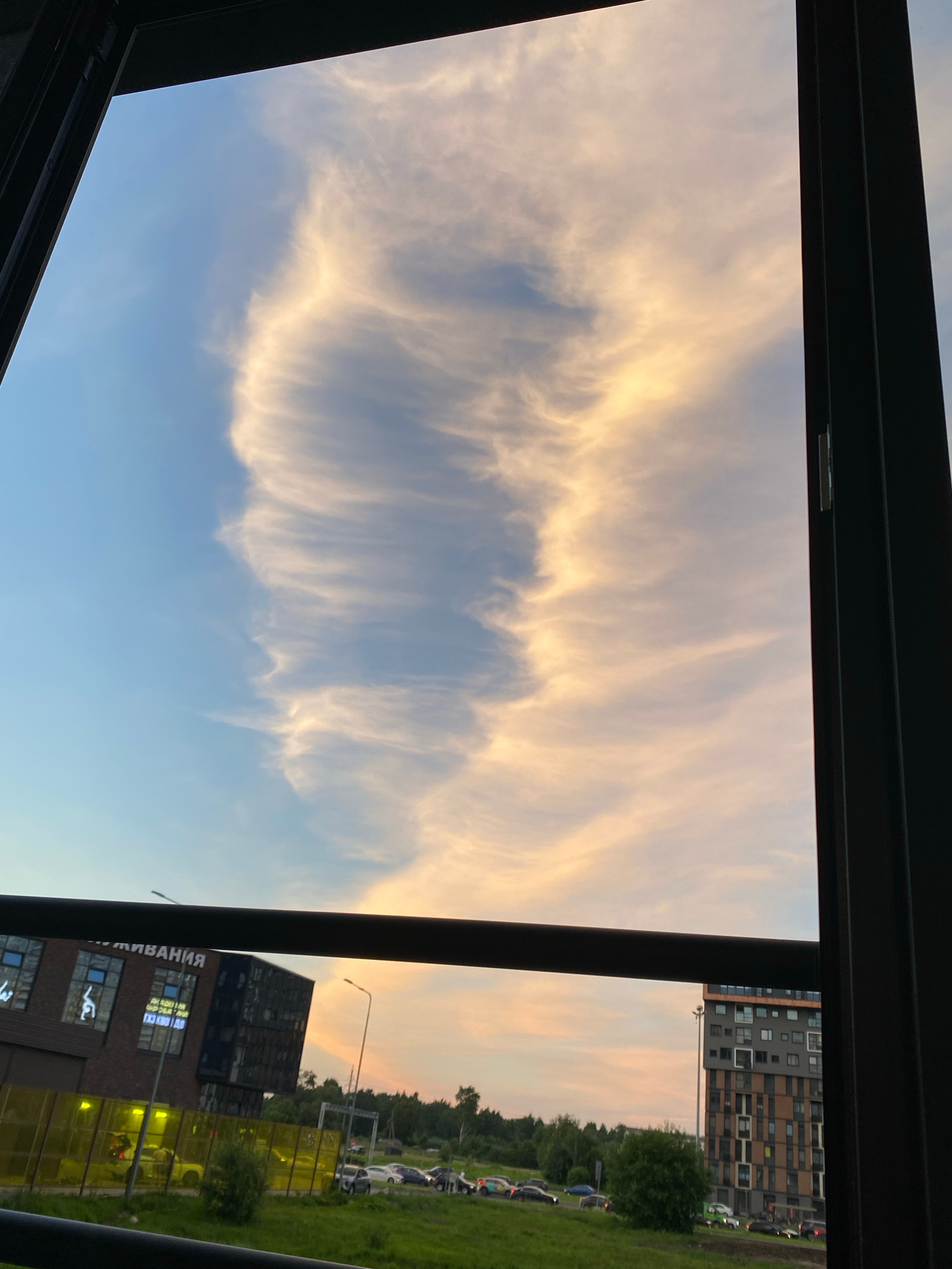 The image captures a view through a window frame, showcasing a sunset over a suburban area. The sky is filled with wispy clouds tinged with orange and yellow hues, contrasting with the clear blue sky. Below the sky, a dark horizontal beam cuts across the frame, separating the sky from the ground level. On the left, a modern, dark-colored building with many windows is visible, with text in Russian on its facade. A yellow barrier runs in front of this building. To the right, a multi-story residential building with a patterned facade stands. The ground level features a grassy area with parked cars and trees. The scene appears to be an urban or suburban landscape, possibly in Saint Petersburg, Russia, given the Cyrillic text on the building. No people are clearly visible in the image.
