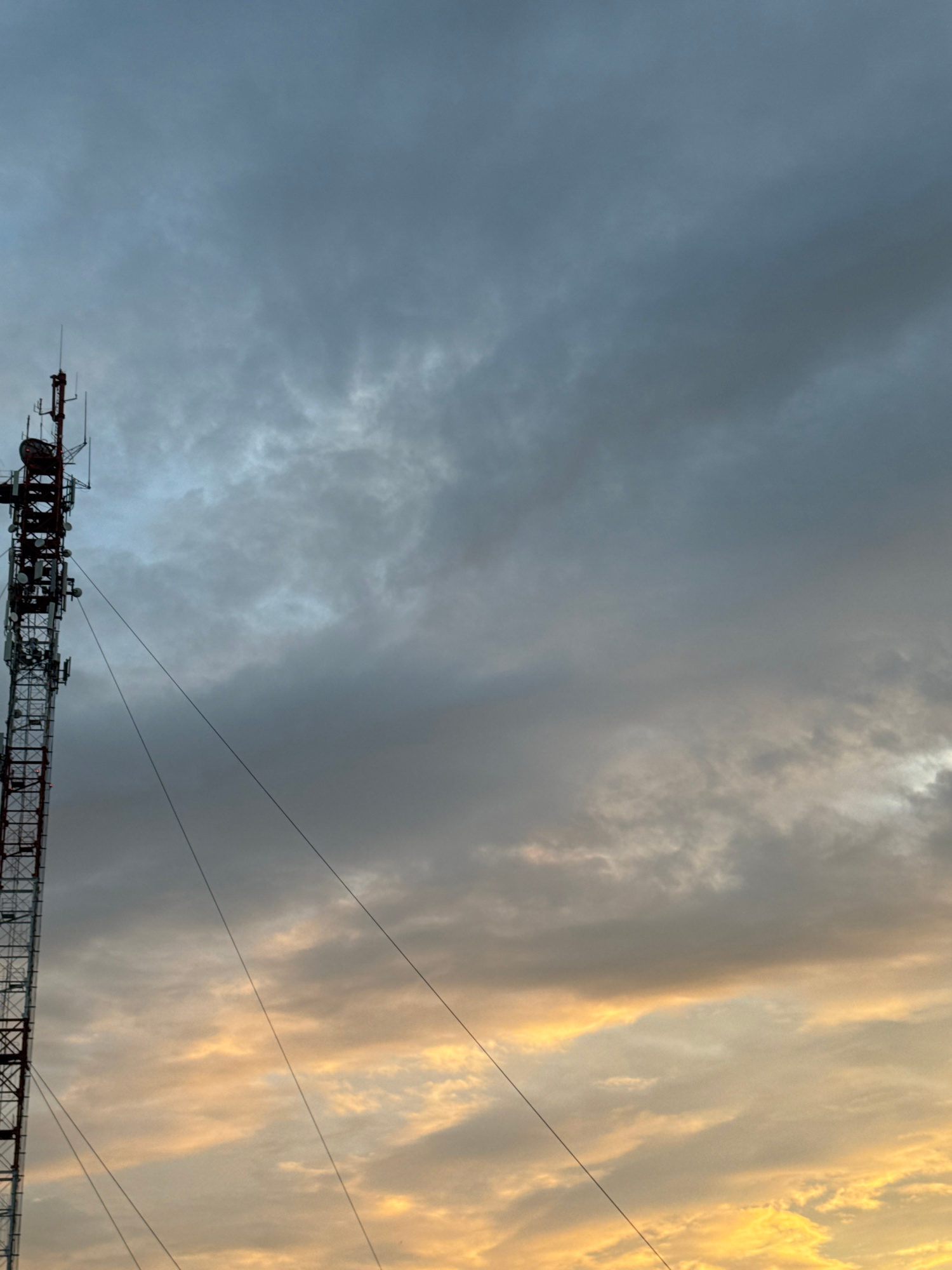 An outdoor, low-angle, vertical shot captures a cell tower against a dramatic sky. The tower, painted red and white, dominates the left side of the frame, with its complex structure of antennas and equipment visible. Several guy wires extend diagonally from the tower, disappearing out of frame. The sky is filled with heavy, grey clouds, but towards the bottom right, the clouds are illuminated by the golden hues of sunset or sunrise, creating a striking contrast. The scene appears to be set in or near the city of Zaokskiy, Russia, given the context of the location. There are no people or any other discernable objects in the immediate vicinity.