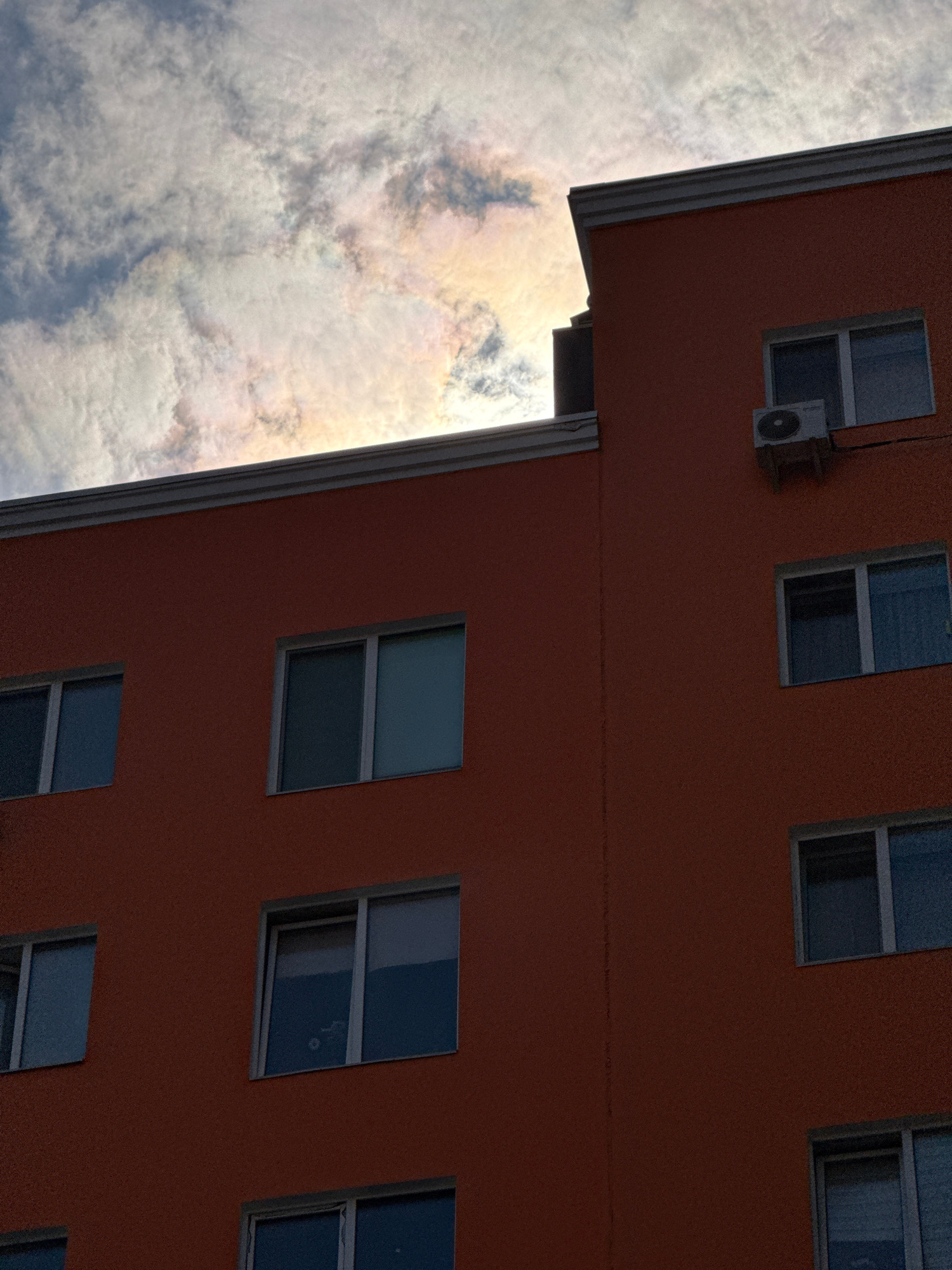 The image shows the side of an orange building with multiple windows and an air conditioning unit. Above the building, the sky is filled with white and grey clouds that are illuminated with iridescent colors, suggesting a phenomenon like iridescence or a sundog. The building's architecture includes a distinct white cornice. The scene appears to be an urban environment in Irpin, Ukraine. There are no people or discernible actions visible in the image.