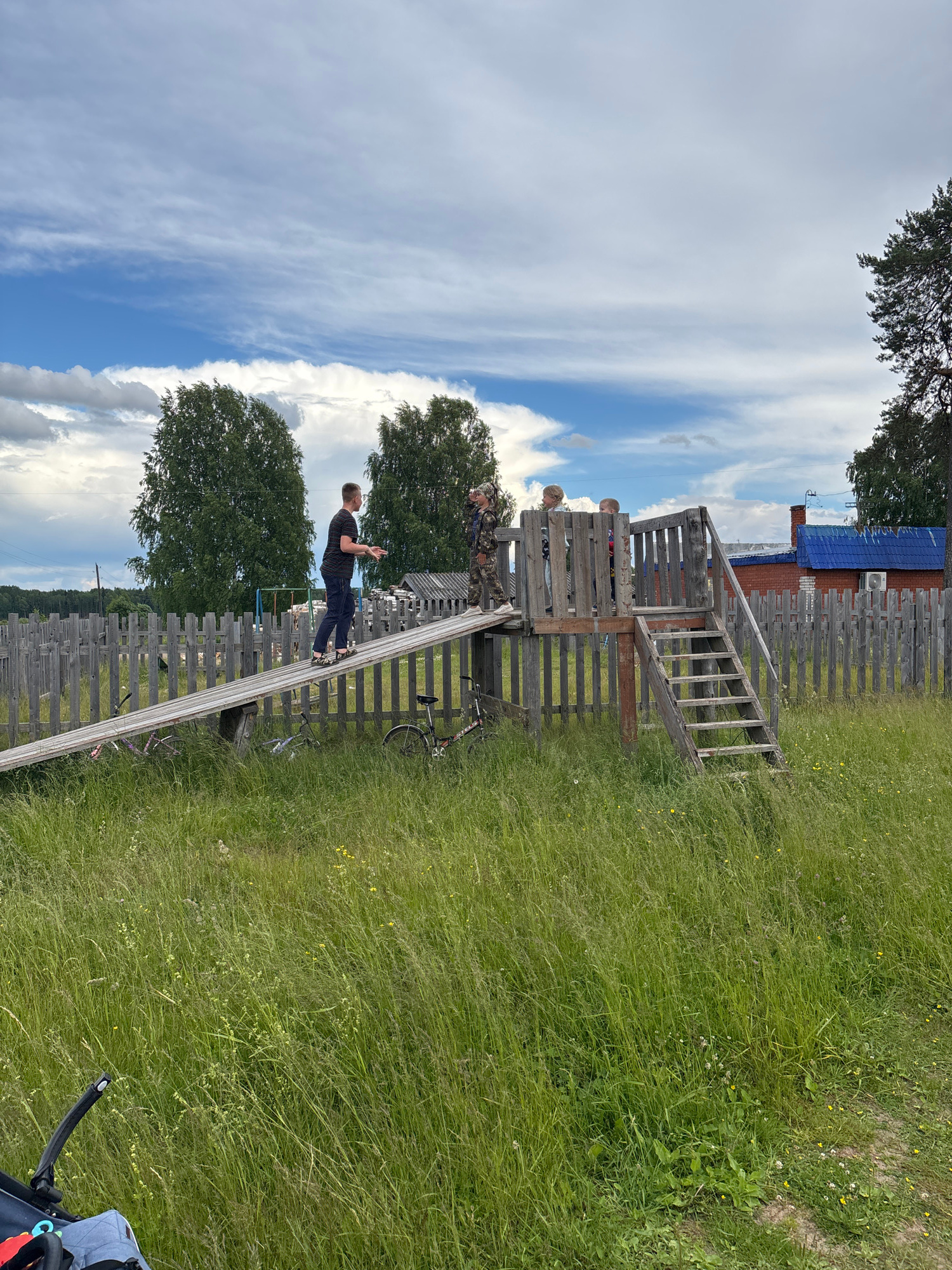 The media file displays a playful scene in a rural setting, likely in Kurgan, Russia. The main subject is a young boy dressed in a black t-shirt and jeans, captured mid-action as he walks down a steep, wooden ramp. He appears to be balancing, with his arms outstretched for stability.
Behind the boy, a rustic wooden structure, possibly a playground or observation deck, is visible. Two other figures, likely children, are standing on this structure. The entire scene is framed by a weathered wooden fence that encircles the grassy area. In the background, a house with a blue roof and a red brick facade can be seen. A bicycle is parked near the wooden structure.
The setting is a sunlit outdoor area with tall, green grass dotted with small yellow flowers. The sky above is a vibrant blue, partially obscured by wispy white clouds, suggesting pleasant weather during daytime. The overall atmosphere is one of carefree childhood activity and simple rural life. There is no visible text in the image.