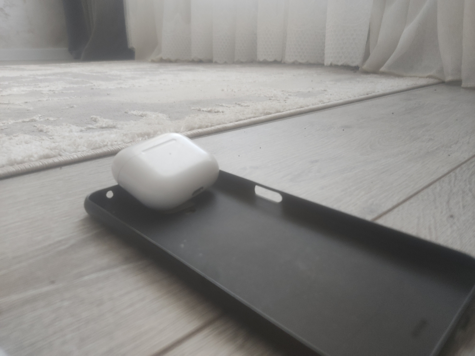 A close-up, low-angle shot shows a white, square-shaped object, possibly a wireless earbud case, attached to the underside of a dark gray shelf. The shelf is positioned horizontally above a light-colored wooden surface, which appears to be a floor or table made of planks. The wooden surface has a visible grain and texture, with some areas appearing slightly worn or dusty. Below the shelf, white, translucent curtains are visible, suggesting an indoor setting. The lighting is soft and diffused, indicating it could be daytime, but there are no direct light sources visible. The focus is on the object and the shelf, with the background slightly blurred. There is no visible text. The scene is simple and domestic, with no people or animals present. The overall impression is one of an everyday object placed in a functional or decorative manner. The context of "Мээнеткеч, Kyrgyzstan" provides a geographical location but offers no specific visual cues within the image itself.