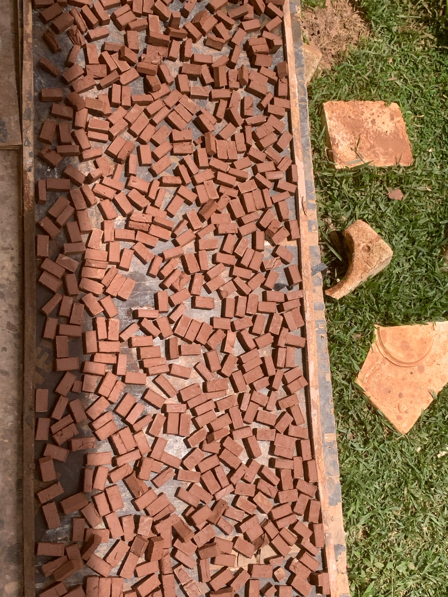 An overhead view captures a collection of small, rectangular, reddish-brown clay bricks spread out on a dark, rectangular surface, likely a drying rack or tray. These bricks are arranged in a seemingly haphazard manner, with some lying individually and others clustered together. The surface they are on appears weathered and possibly made of wood or metal, with visible grain or texture.
To the right of the tray, a patch of green grass is visible, suggesting an outdoor setting. On the grass, three larger, irregularly shaped, light orange-brown objects are present. One is a flat, square piece, appearing to be a broken shard of terracotta. Another is a curved, hollowed-out object, possibly a fragment of a broken pot or roof tile, with a distinct opening. The third is a larger, flat, square piece, also with a terracotta-like texture, lying slightly angled.
The overall scene appears to be a location within Lafia, Nigeria, where these small bricks are being sun-dried, likely for a craft or construction purpose. No people or animals are depicted in the image. The lighting suggests a sunny day, casting faint shadows from the bricks onto the surface.