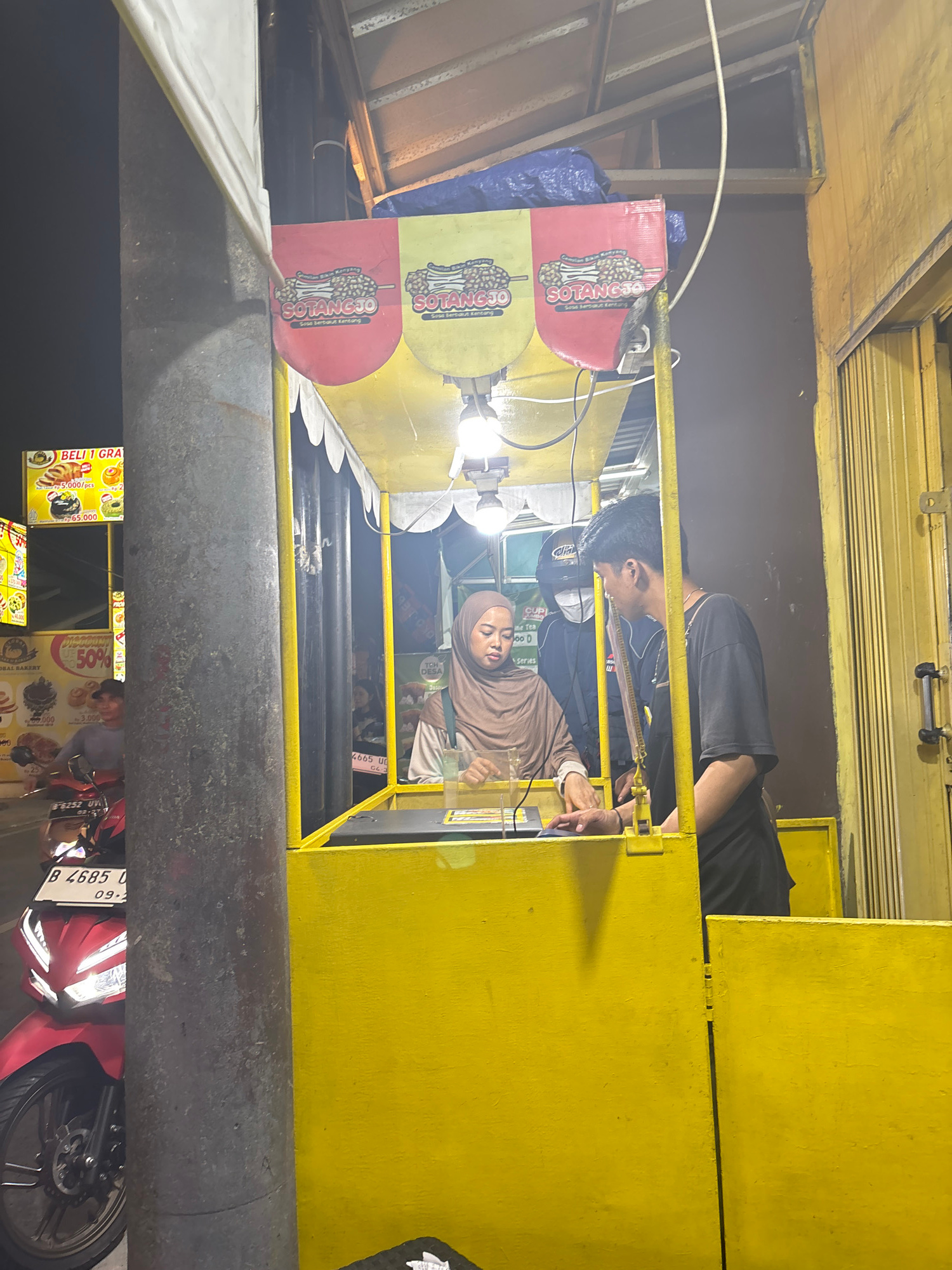 This media file shows a street-level view of a food stall in North Jakarta, Indonesia, at night. The stall is brightly lit with yellow paint and features a yellow metal frame. Two individuals are visible behind the counter. One is a young man wearing a black t-shirt and a face mask, leaning over the counter. The other is a young woman wearing a brown hijab and a light brown top, also behind the counter.
On the stall, there are signs with text in Indonesian. One sign advertises "SOTANGJO" with an image of what appears to be food on a stick, and mentions "Sosis berbalut Kentang" (sausage wrapped in potato). Other signs display prices such as "5.000/pcs" and "65.000". There's also a sign for "BAL BAKERY" with a "50%" discount and prices for baked goods. Further signs show "CUPI" and "Fine Tea". License plates are visible, including "B 4685 UG" and "B 4685 O".
In the foreground, a large, weathered metal pipe is prominently featured, partially obscuring the view of the stall and a red motorcycle parked nearby. The overall scene suggests a busy, late-night food stall operation. The lighting indicates it is nighttime, and there are no clear indications of weather conditions.