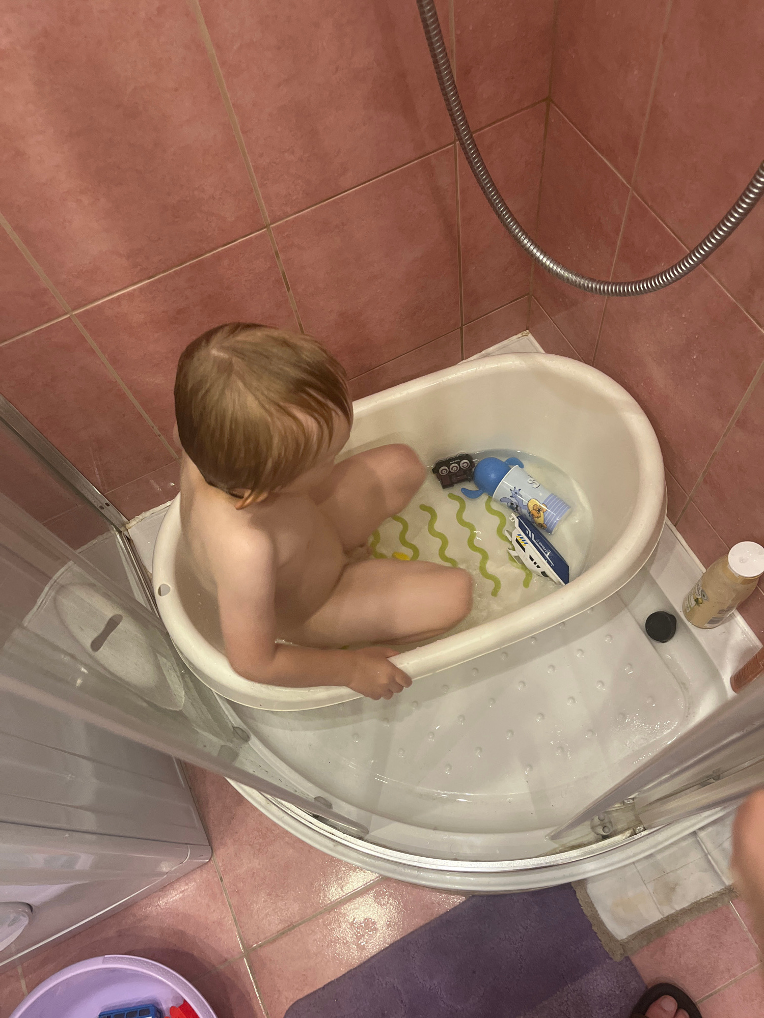 In a bathroom, a young child with blonde hair is sitting in a white bathtub, taking a bath. The child is partially submerged in water and their legs are visible. There are two bath toys in the tub: a blue and white toy boat and a blue cylindrical toy with a character on it, possibly a duck. The bathtub is inside a shower stall with a transparent glass door. The floor is tiled with reddish-brown tiles, and a gray hose is visible on the left side of the frame. A white bottle with a pump dispenser is also present in the shower stall. The overall impression is a domestic scene of a child's bath time. No specific time of day or weather can be determined.