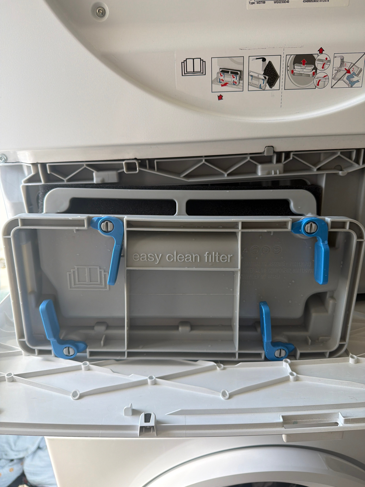 The image shows an open washing machine filter compartment. The compartment is gray plastic with two blue levers on the left side for opening and closing it. A black filter is visible inside the compartment. Text on the inside of the compartment reads "easy clean filter". Above the filter compartment, on the white exterior of the washing machine, are instructions for cleaning the filter, depicted in a series of diagrams. These diagrams show the filter being removed, cleaned under running water, and then reinserted. The washing machine appears to be located indoors, likely in a domestic setting. There are no people visible in the image, and no specific location information is provided.