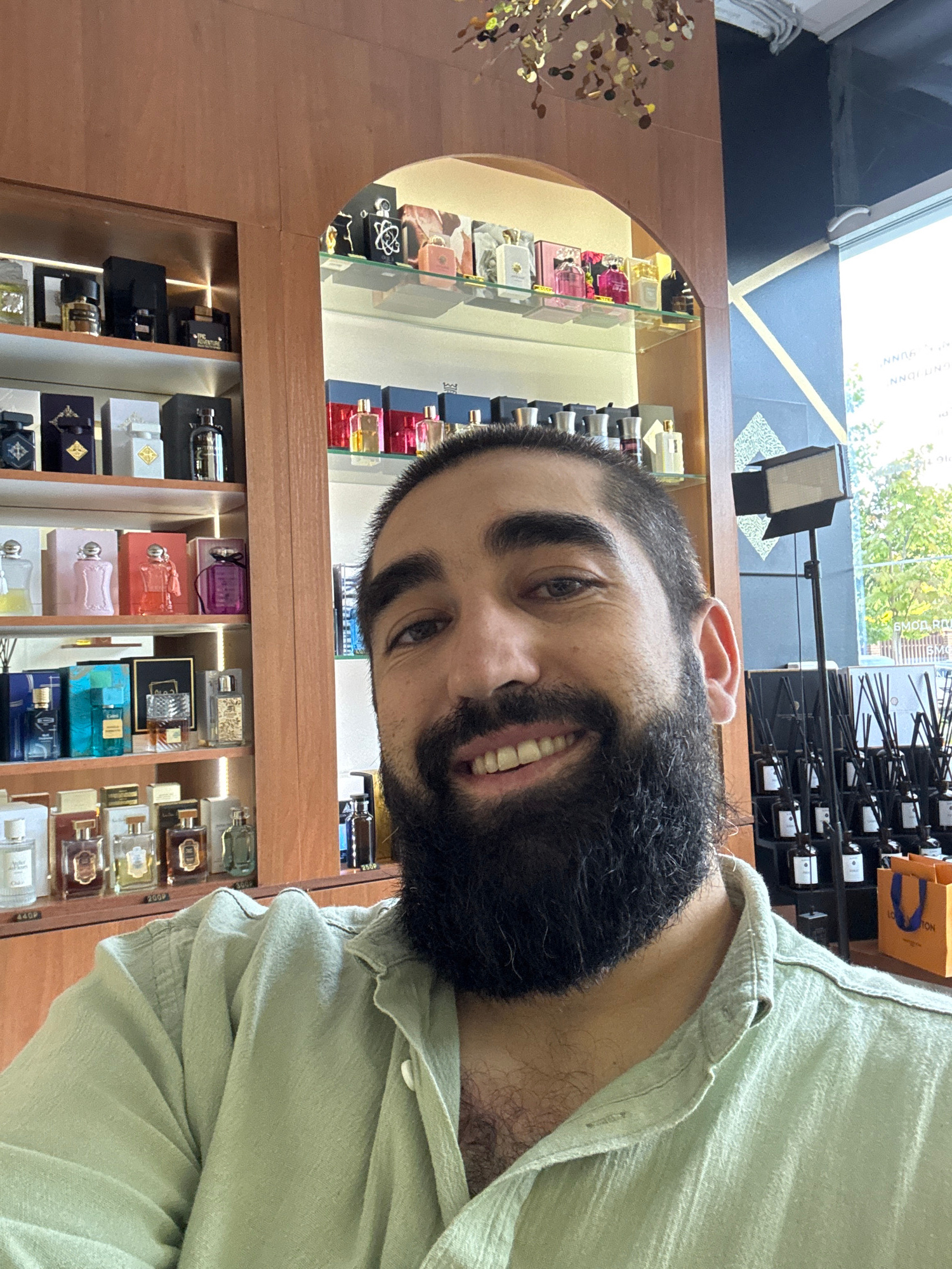 A man with a full beard and dark hair smiles at the camera, taking a selfie. He is wearing a light green collared shirt. Behind him, on shelves, are numerous bottles of perfume and diffusers. The shelves are made of light-colored wood and are illuminated. To the right, a window shows trees and a sky. The overall setting appears to be a perfume shop. There is no information in the image to indicate it is located in Krasnodar, Russia.