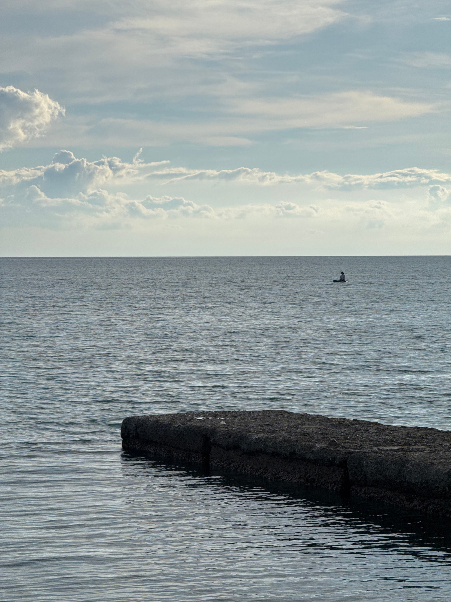 This image depicts a calm body of water, likely the Black Sea, with a wide expanse of water stretching to the horizon. In the foreground, a dark, rectangular concrete structure juts out into the water, partially submerged. The surface of the water is rippled, suggesting a gentle movement. In the distance, a single person is visible on a paddleboard, silhouetted against the horizon. The sky above is partly cloudy, with soft, diffused light creating a serene atmosphere. The scene evokes a sense of tranquility and solitude. The location is not specified in the image, but given the context of Sochi, Russia, this could be a typical coastal view from its shoreline.