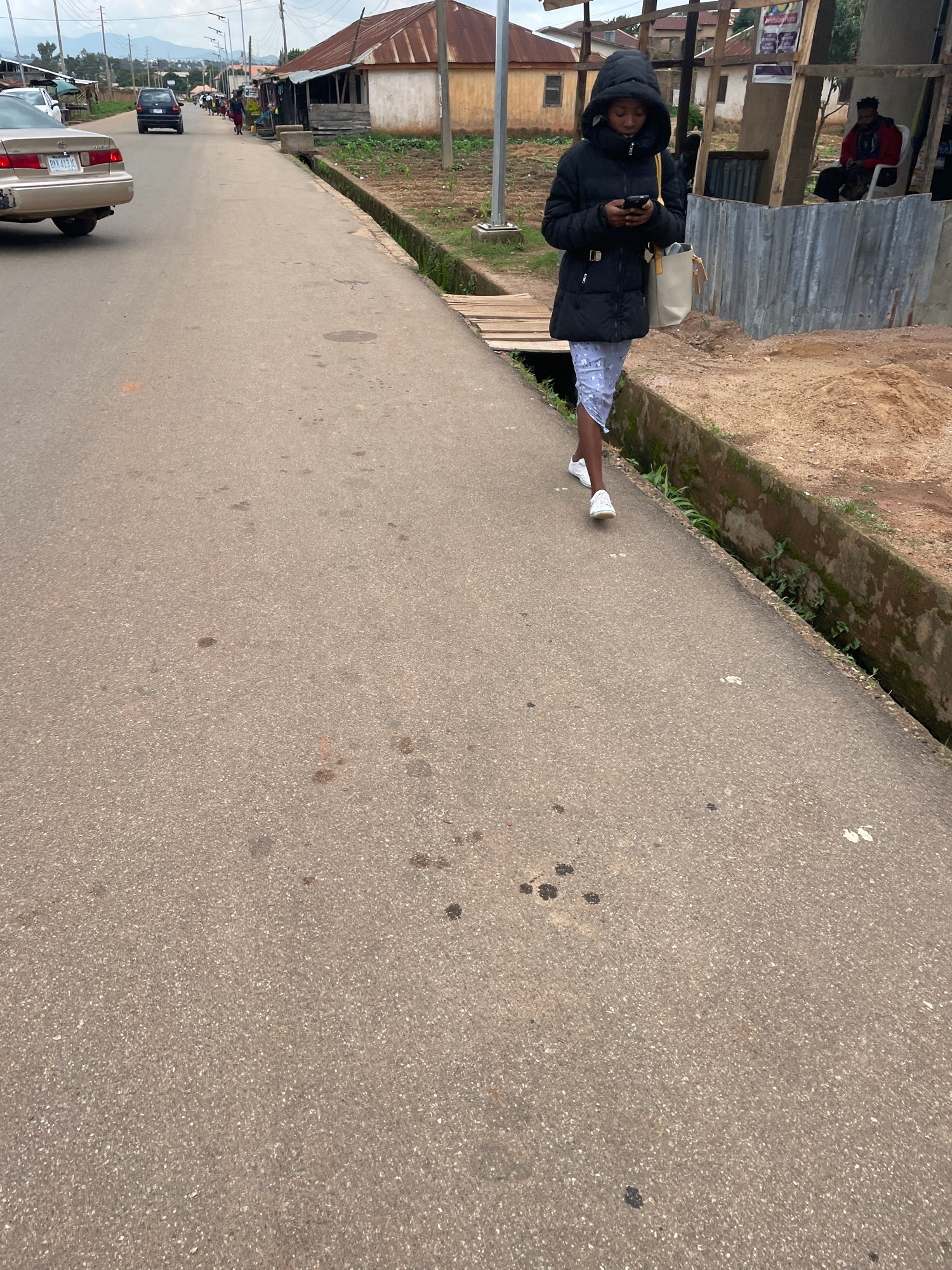 In Barikin Jirga Summa, Nigeria, a young woman wearing a black puffer jacket and a light-colored patterned skirt walks on a paved road, looking at her phone. She carries a light-colored tote bag over her shoulder. Beside the road is a drainage ditch and beyond that, a dusty area with some vegetation and buildings. Several cars are visible on the road in the background, along with other pedestrians. The environment appears to be a typical street scene in a Nigerian town.