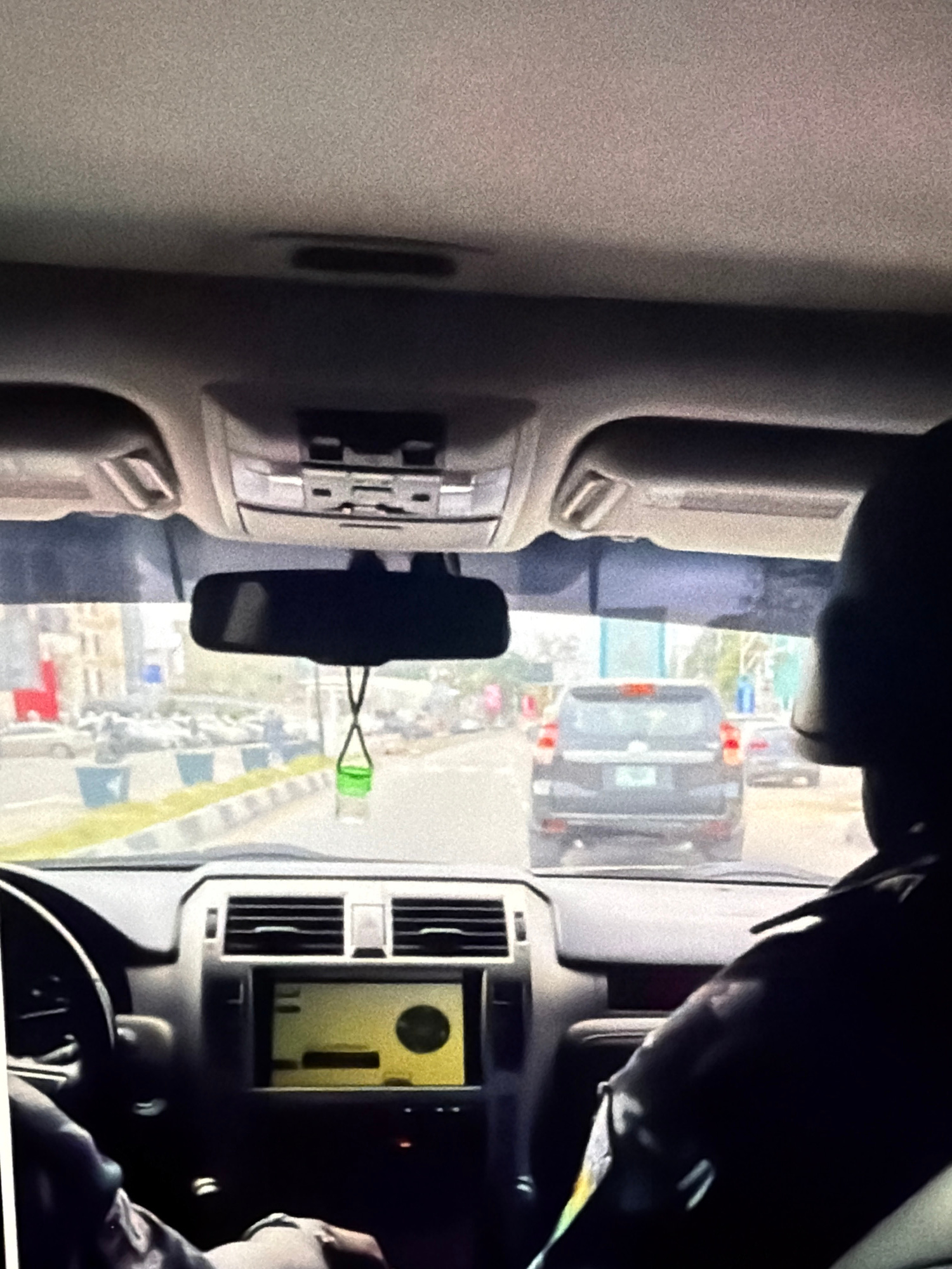 The image is a first-person view from inside a vehicle, looking forward through the windshield. The vehicle is on a paved road in what appears to be a city. In the foreground, the dashboard of the vehicle is visible, including the steering wheel on the left and a multimedia screen in the center. A person's arm wearing a dark jacket is visible on the right side of the frame, suggesting they are the driver or a passenger in the front.
Outside the vehicle, traffic is present. A dark-colored SUV is directly ahead, followed by other vehicles. The buildings and infrastructure suggest an urban environment. A green air freshener is hanging from the rearview mirror. The road has lane markings and what appear to be traffic signs or barriers on the side. The lighting indicates it is daytime.