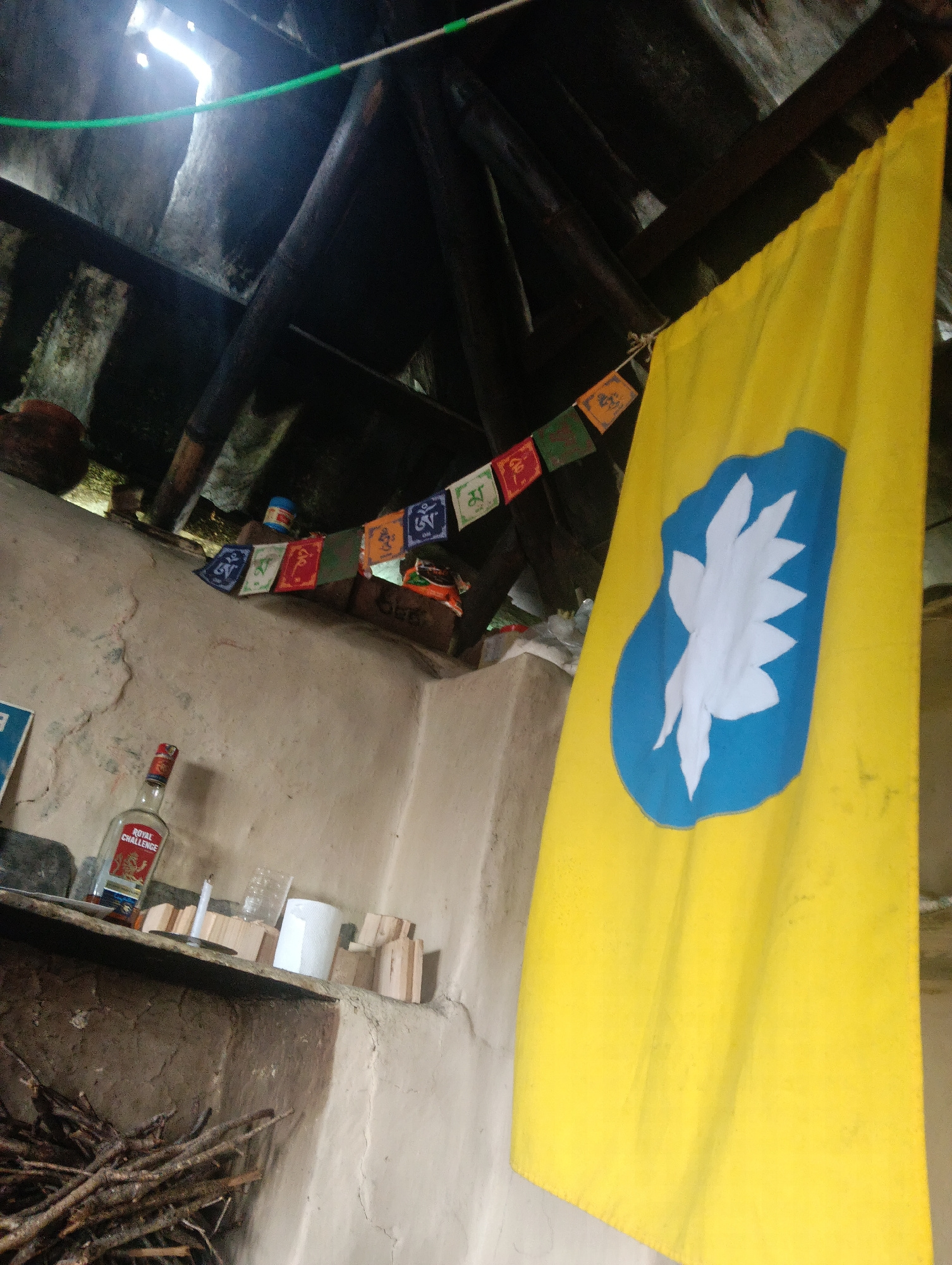 The image depicts an interior scene within a rustic structure, likely a dwelling or a hut, with exposed wooden beams and a rough, textured ceiling suggesting a rural or underdeveloped setting. A large, bright yellow flag with a blue oval containing a white, star-like emblem dominates the right side of the frame. Suspended from a string above and to the left of the flag are a series of small, colorful prayer flags, each bearing Tibetan script.
On a shelf in the mid-ground, a bottle of "Royal Challenge" whisky stands next to a candle, a plastic container, a roll of white paper, and several small wooden blocks. To the left of the shelf, a pile of firewood is visible. The wall surfaces are rough and uneven, painted in a pale, earthy tone. Natural light streams in from an opening in the ceiling, creating bright highlights and shadows. The overall impression is one of simplicity and a connection to traditional or spiritual practices, potentially in a remote location. The mention of Haini, India, suggests a geographical context, but the image itself does not contain specific identifiers of this location.