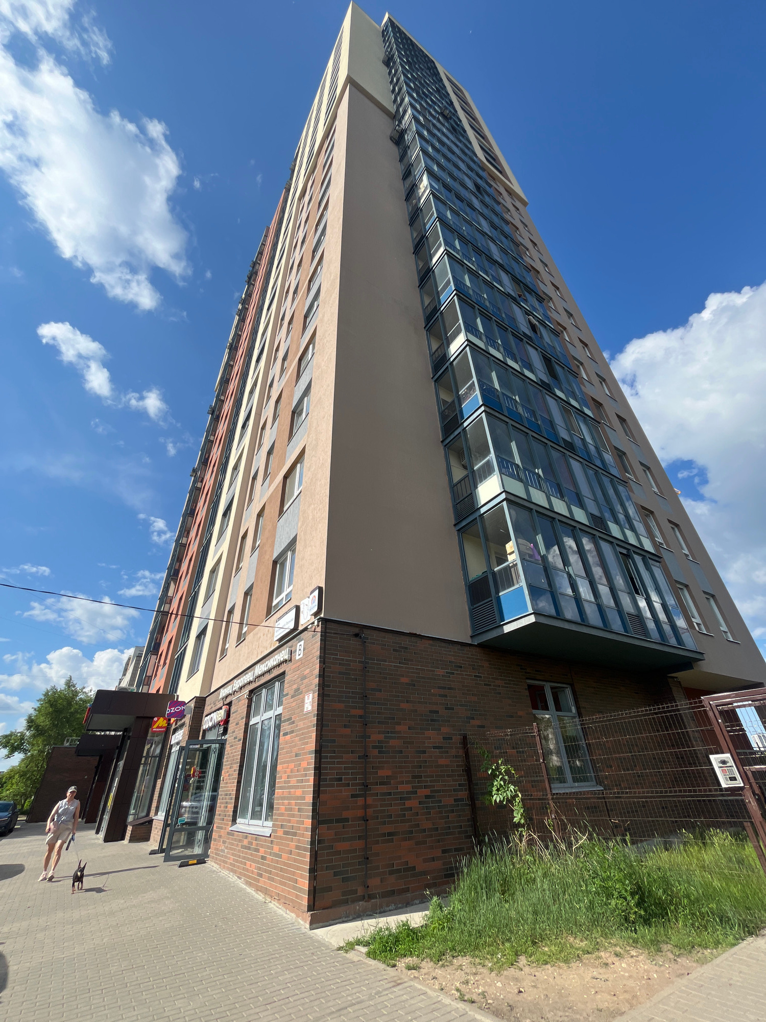 A low-angle, full-shot photograph captures a tall, modern apartment building on a sunny day in Perm, Russia. The building's facade is a mix of light tan, grey, and vertical bands of orange and yellow. The lower portion of the building is clad in dark red brick, featuring large glass-fronted shops on the ground floor. To the left, a woman wearing a light-colored shirt and shorts walks her small black dog along the paved sidewalk. The sky is a vibrant blue with scattered white clouds. A metal fence encloses an area with overgrown green grass and weeds in front of the building's brick base. The image appears to be taken on a street in a residential or mixed-use urban area.