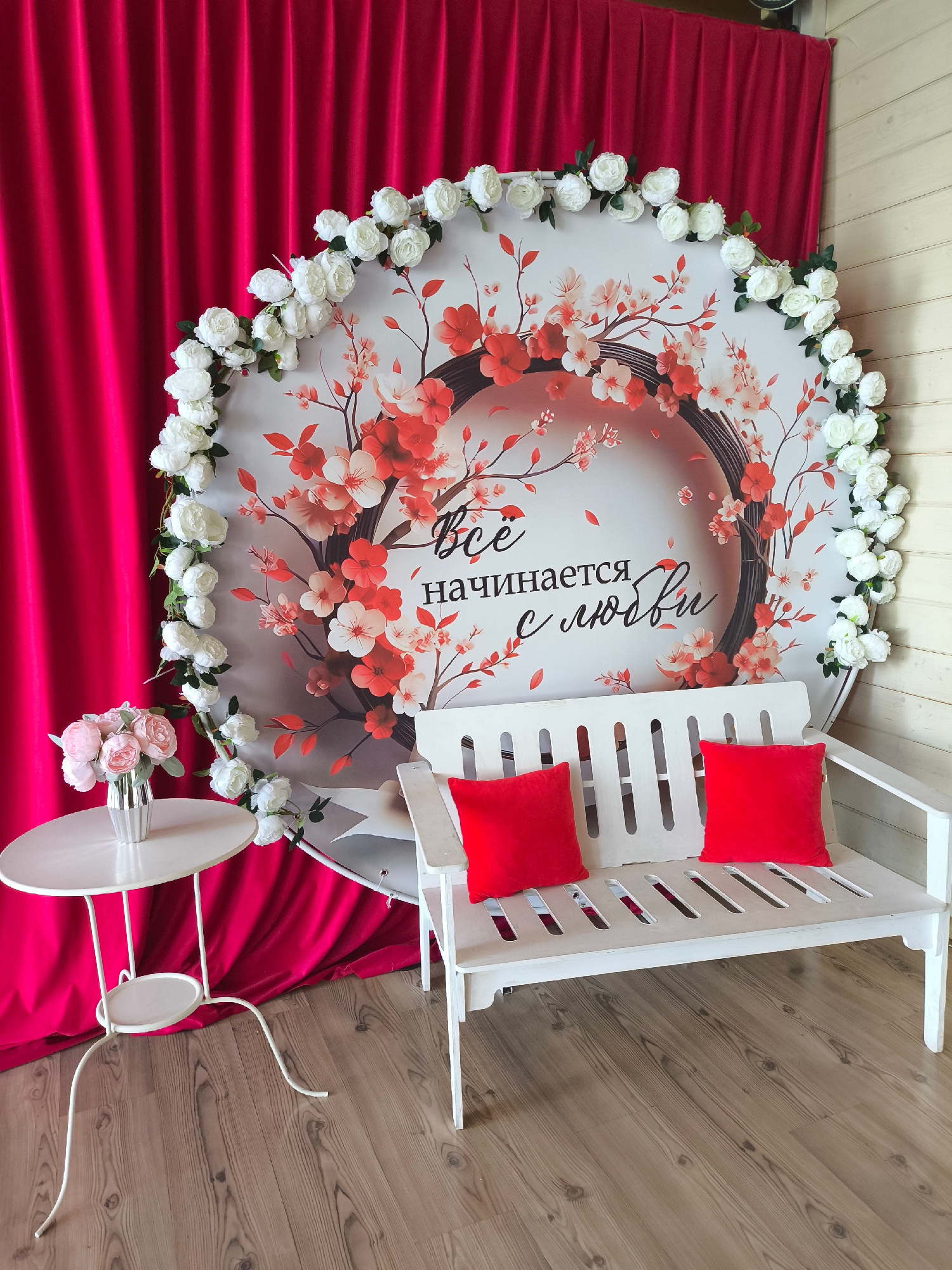This is a forensic, matter-of-fact description of a scene located in Taldom, Russia. The scene depicts a photo zone with a white wooden bench in the foreground, adorned with two red square cushions. To the left of the bench is a small, white, round side table with a vase containing a bouquet of pink and white flowers. Behind the bench and table, a large, circular backdrop features a floral design with cherry blossoms and the Russian text "Всё начинается с любви" (Everything begins with love). White artificial flowers are arranged in a garland along the edge of the backdrop. To the left of the backdrop, a vibrant fuchsia curtain is draped. The floor is made of light-colored wooden planks. The background wall to the right of the backdrop appears to be made of light-colored wooden planks as well. No people are present in the scene. The lighting appears to be natural or artificial, creating a bright and clear image.