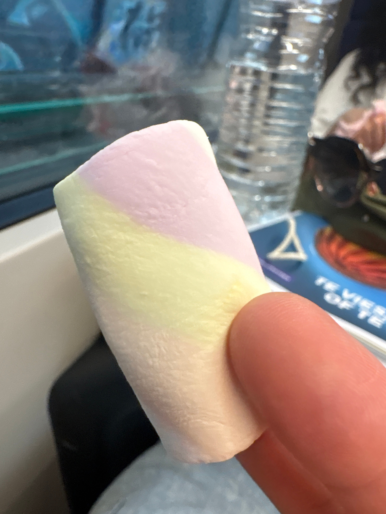 A close-up, high-angle shot captures a person's fingertip holding a tri-colored marshmallow. The marshmallow exhibits swirls of pink, yellow, and white. In the background, a blurred water bottle and sunglasses are visible, alongside a magazine with the words "TE VIE OF TE" printed on it. The scene appears to be set on a mode of transportation, possibly a train, given the window and reflections suggesting movement. The overall setting is ambiguous and does not provide any specific indicators of being in Delft, Netherlands.