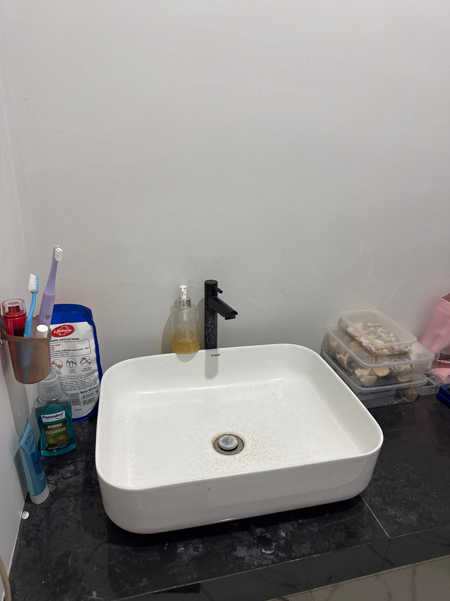 The image depicts a bathroom sink area. A white, rectangular sink is positioned on a dark, speckled countertop. A black faucet with a single handle is mounted behind the sink. To the left of the sink, a metal holder contains two toothbrushes and a red-capped item, possibly lipstick or a similar cosmetic. A bottle of Pepsodent mouthwash and a tube of Ponds toothpaste are also visible on the countertop. Behind these items, a large blue and white bag with text in an Asian language is propped up. To the right of the sink, a clear pump dispenser with yellow liquid, likely hand soap, is present. Further to the right, stacked clear plastic containers filled with seashells are on the countertop. The background consists of a plain white wall with subtle cracks. The scene is consistent with a bathroom in a residential setting in Sukawati, Indonesia, given the context provided.