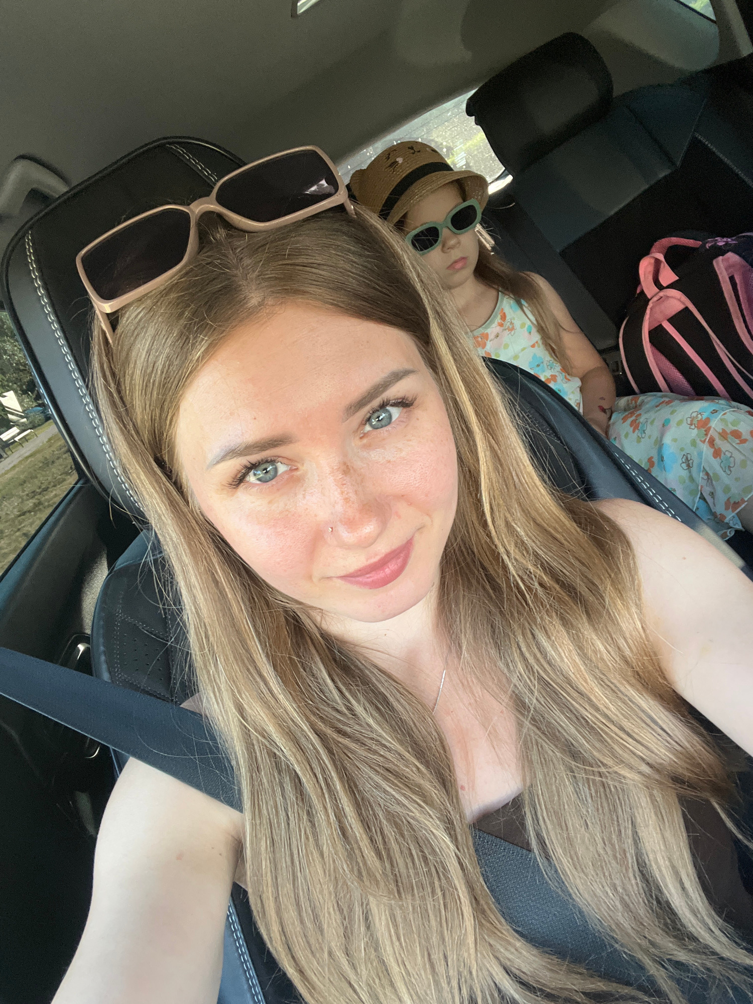 This is a selfie taken from inside a car, showing a woman with blonde hair and blue eyes, wearing sunglasses on her head. She has freckles and a nose piercing. In the background, a young girl with blonde hair is visible, wearing a straw hat and sunglasses. She is seated in the car, facing slightly away from the camera. A backpack with pink accents is also visible in the background, behind the girl. The image appears to be taken during daytime, with natural light entering the car. The setting is likely a road or parking lot, with trees and greenery visible through the car window on the left. There are no identifiable landmarks or specific features that would indicate the location as Stepnogorsk city administration, Kazakhstan.