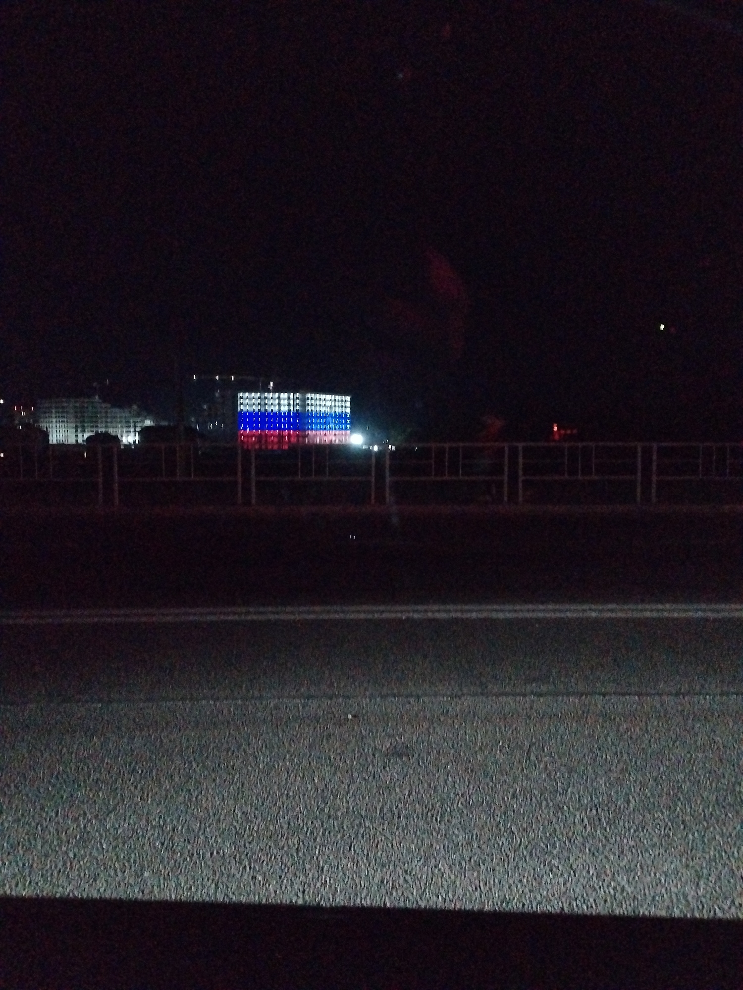 The image captures a nighttime scene from a slightly elevated perspective, looking across a dark road towards a building illuminated with the colors of the Russian flag. The building, which appears to be a large structure, has its windows lit in horizontal bands of white, blue, and red, forming a large-scale representation of the national flag. In the foreground, a dark asphalt road is visible with white lane markings. Beyond the road, a metal railing separates the viewer from the area where the building is situated. The background is predominantly dark, with some faint lights suggesting other structures or distant illumination. The overall atmosphere is one of a city at night, with a prominent display of national symbolism.