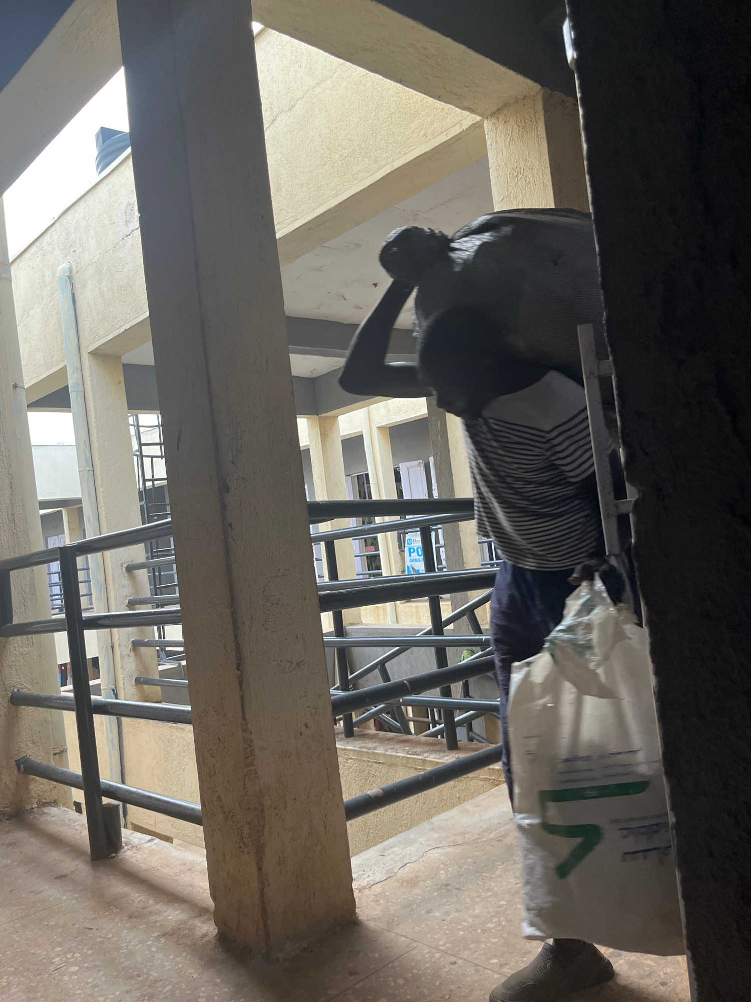 An unidentified man carries a large, dark, irregularly shaped bundle on his shoulders and a white plastic bag in his right hand as he walks down a concrete staircase. He wears a striped t-shirt and dark pants. The staircase is outdoors, with railings on both sides. The building appears to be made of concrete and is multi-storied, with balconies visible in the background. The man is walking down the stairs, his face obscured by the bundle on his shoulders. The image suggests an urban environment, possibly a residential or commercial building complex, with the characteristic architecture of Jos, Nigeria. The lighting indicates it is daytime.