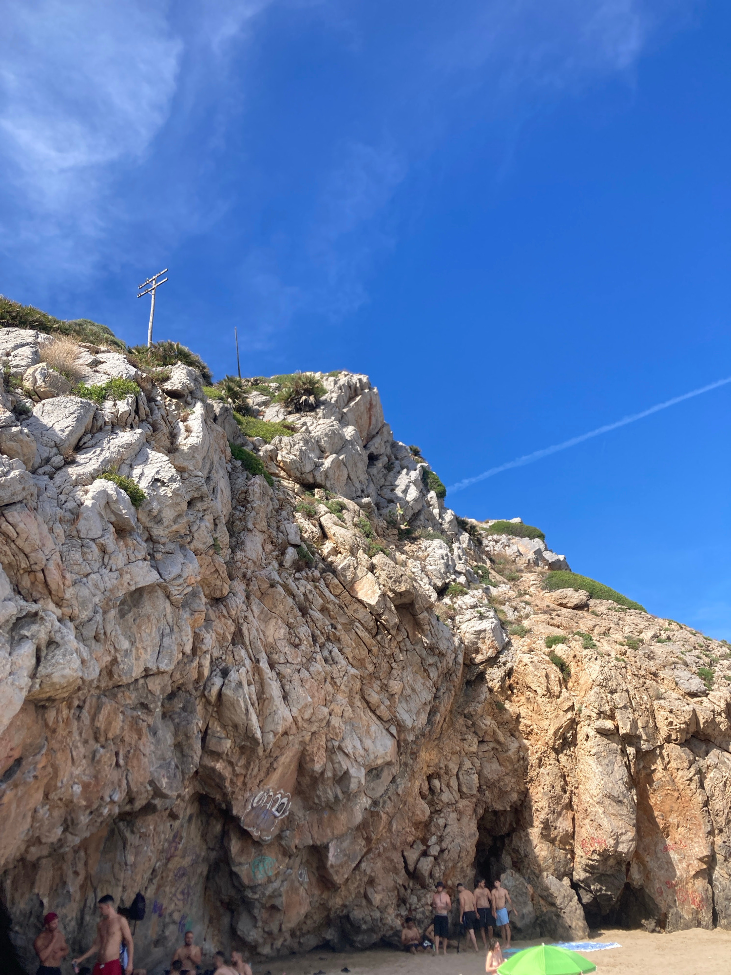 A high-angle, medium shot captures a rocky cliff face on a beach in Port De La Ginesta, Spain. The cliff, composed of layered, rough-textured rock in shades of tan, orange, and gray, dominates the left and center of the frame. Sparse green vegetation grows from crevices in the rock. At the top of the cliff, a wooden utility pole with two crossbars stands against a bright blue sky filled with wispy white clouds and a faint contrail. The lower right portion of the image reveals a sandy beach with a few people gathered near the base of the cliff, some of whom are partially obscured by the shadow of an overhang. Several individuals are standing and conversing, while others appear to be sitting or relaxing. A bright green umbrella is partially visible in the foreground, suggesting a recreational setting. Graffiti in white, red, and green is present on the lower sections of the cliff face.