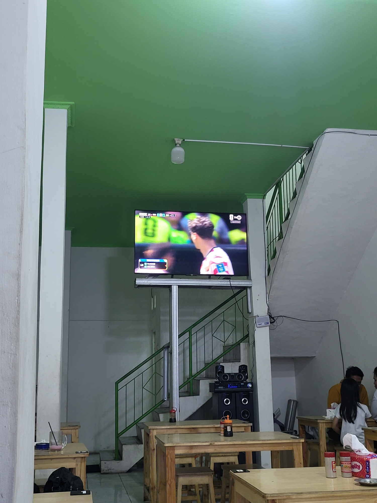 This is an indoor scene, likely a restaurant or cafe, with a green ceiling. A large television screen is mounted on a stand and displays a soccer match with player statistics and a logo for "TVRI LANGSUNG". There are wooden tables and chairs set up. A set of stairs with a green railing is visible, leading to an upper level. On the right side of the image, three people are seated at a table, with two appearing to be interacting. One person is wearing a yellow shirt, and another is wearing a white shirt. A tissue box is on the table in front of them. A sound system with speakers is placed near the base of the stairs. Various objects, including cups, a phone, and a bottle of sauce, are present on the tables. The flooring appears to be tiled. The overall impression is a casual, communal space designed for viewing entertainment while dining.