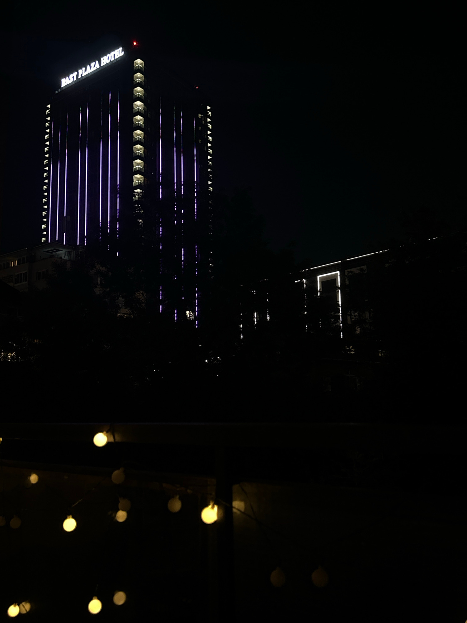 The image shows a nighttime view of the East Plaza Hotel in Sofia, Bulgaria, with its facade illuminated by purple vertical lights. The hotel is a tall, modern building with numerous floors and balconies. To the right of the hotel, another building is visible, also illuminated by lights. In the foreground, a string of yellow lights is blurred, suggesting they are close to the camera and out of focus. The overall scene is dark, with the exception of the illuminated buildings and the foreground lights.