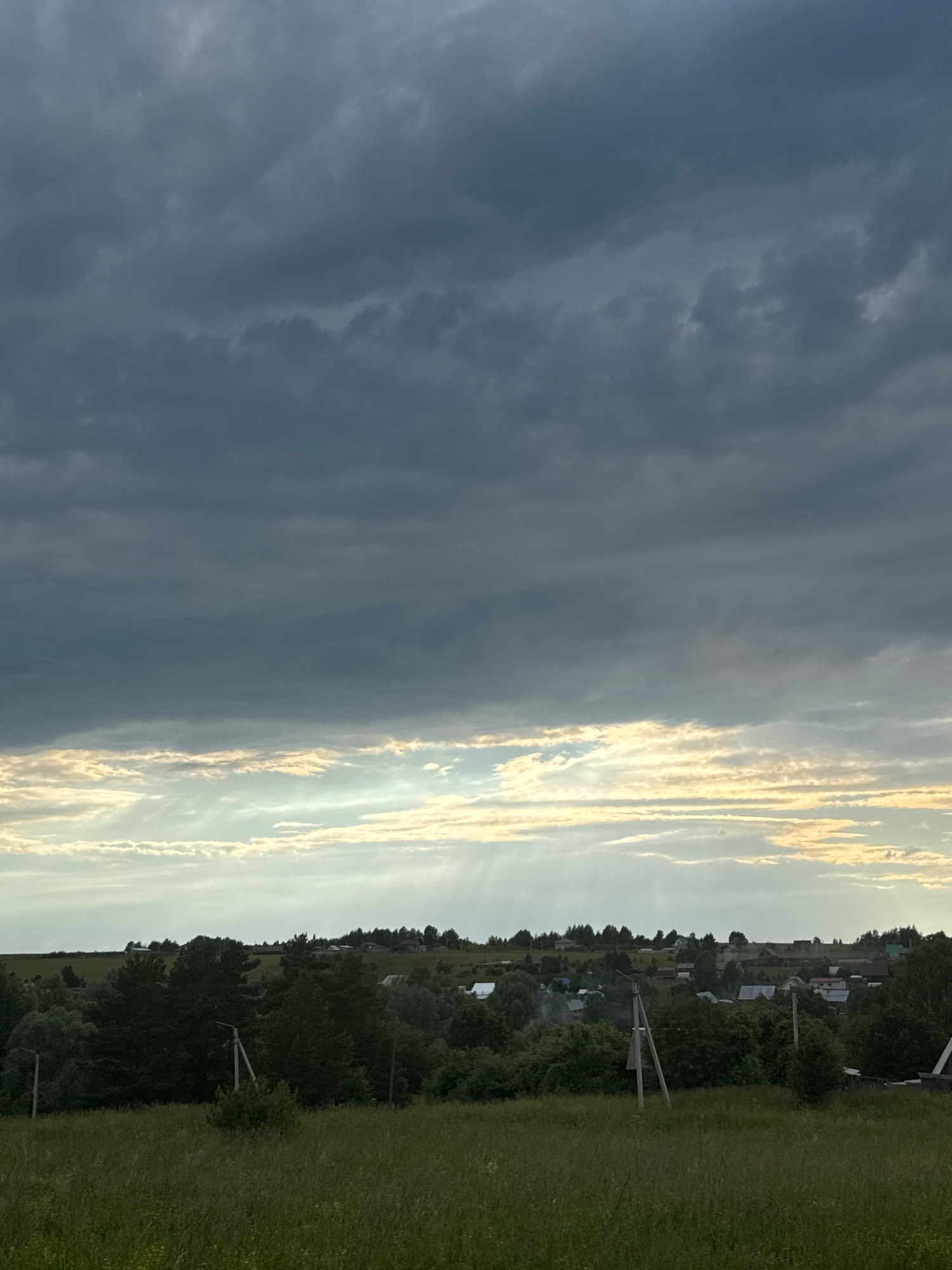 This image depicts a rural landscape under a dramatic sky. Dark, heavy clouds dominate the upper portion of the frame, with lighter, stratiform clouds forming a band near the horizon, pierced by visible sunbeams. Below the sky, a dense line of green trees and foliage stretches across the midground, obscuring the lower parts of a village. Several structures with visible roofs can be seen peeking through the trees and on the distant rise, suggesting a settlement within the Malyi Bukor area of Russia. Two utility poles with wires are prominent in the foreground and midground. The immediate foreground is a field of tall, green grass. There are no people or animals visible in the scene.