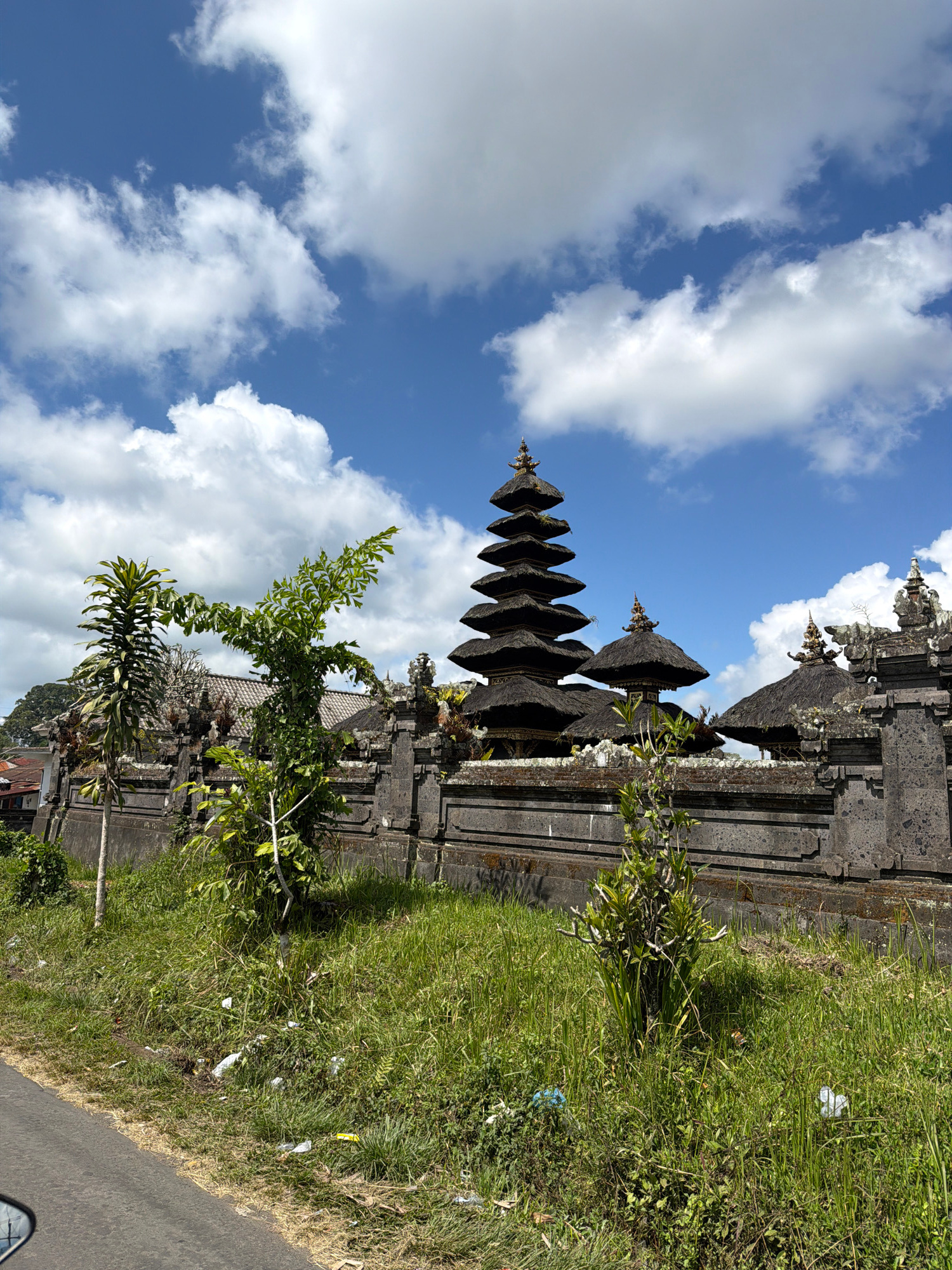 This image depicts a traditional Balinese temple with multiple tiered roofs, known as Meru, set against a bright blue sky dotted with white, fluffy clouds. The temple complex is enclosed by a stone wall with decorative elements. Lush green grass and foliage grow in front of the wall, with a paved road visible in the foreground. The scene is likely located in or around Baturiti, Indonesia, known for its numerous temples and verdant landscapes. There are no people visible in the image.