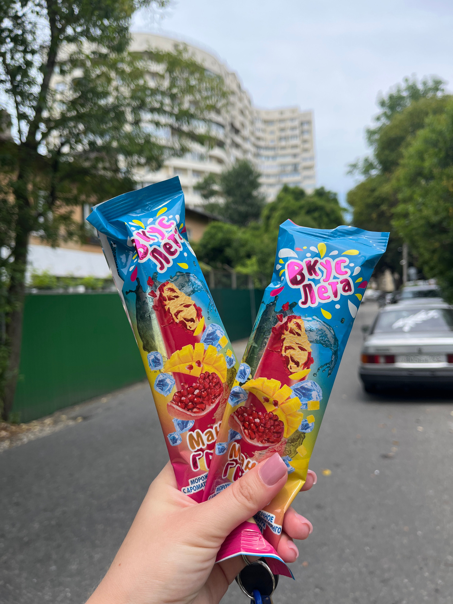 A person's hand holds two popsicle wrappers with blue, pink, and yellow designs depicting ice cubes, water splashes, and fruit slices of pomegranate and mango. The text on the wrappers reads "Вкус Лета" (Taste of Summer) in stylized pink and yellow lettering. Below this, in smaller text, it says "Манго Гранат" (Mango Pomegranate) and "МОРОЖЕНОЕ" (Ice Cream). The background is slightly blurred, showing trees, a building, and a white car on a paved street. The scene appears to be outdoors in a city.