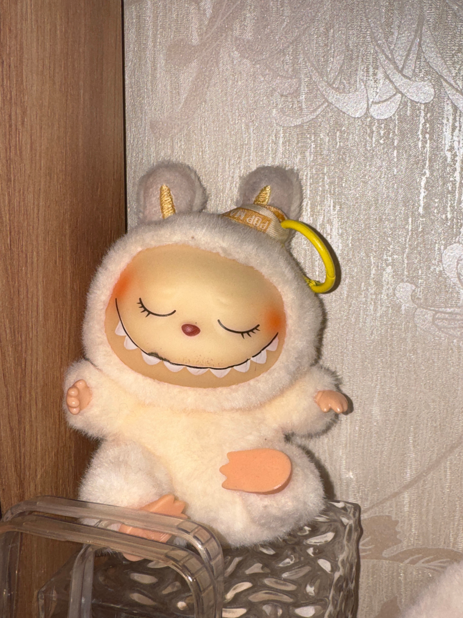 A stuffed toy resembling a fluffy, white creature with a round, yellow face is lying on its side on a wooden surface. The toy has small, peach-colored ears, hands, and feet. Its eyes are closed, and its mouth is depicted as a wide, happy grin with white, tooth-like shapes. A blush of orange is visible on its cheeks. A yellow loop is attached to the top of its head, and a fabric tag with gold lettering is visible on its neck.
Behind the toy, a textured, light-colored wallpaper with a subtle floral pattern can be seen. To the right of the toy, a transparent plastic object with a patterned interior is partially visible. The overall scene suggests a domestic setting, possibly a room. No specific time of day or weather conditions are discernible. The location context is Nizhny Novgorod, Russia. The visible text on the tag appears to read "POP M".