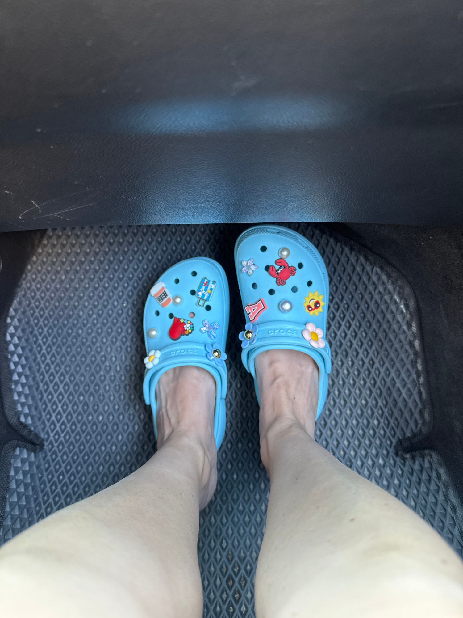 This is a first-person, top-down view of a person's feet wearing light blue Crocs on a textured, dark grey floor mat. The Crocs are adorned with various Jibbitz charms, including pearls, a red lobster, a pill bottle, a sun with a face, a heart with a bandage, flowers, and a butterfly. The surrounding environment appears to be the interior of a vehicle, suggested by the dark, textured material at the top of the frame, which could be a dashboard or car interior panel. The floor mat has a diamond-shaped pattern. The lighting suggests an indoor or shaded outdoor setting. There is no indication of the city of Boreti, Montenegro, in the image itself.