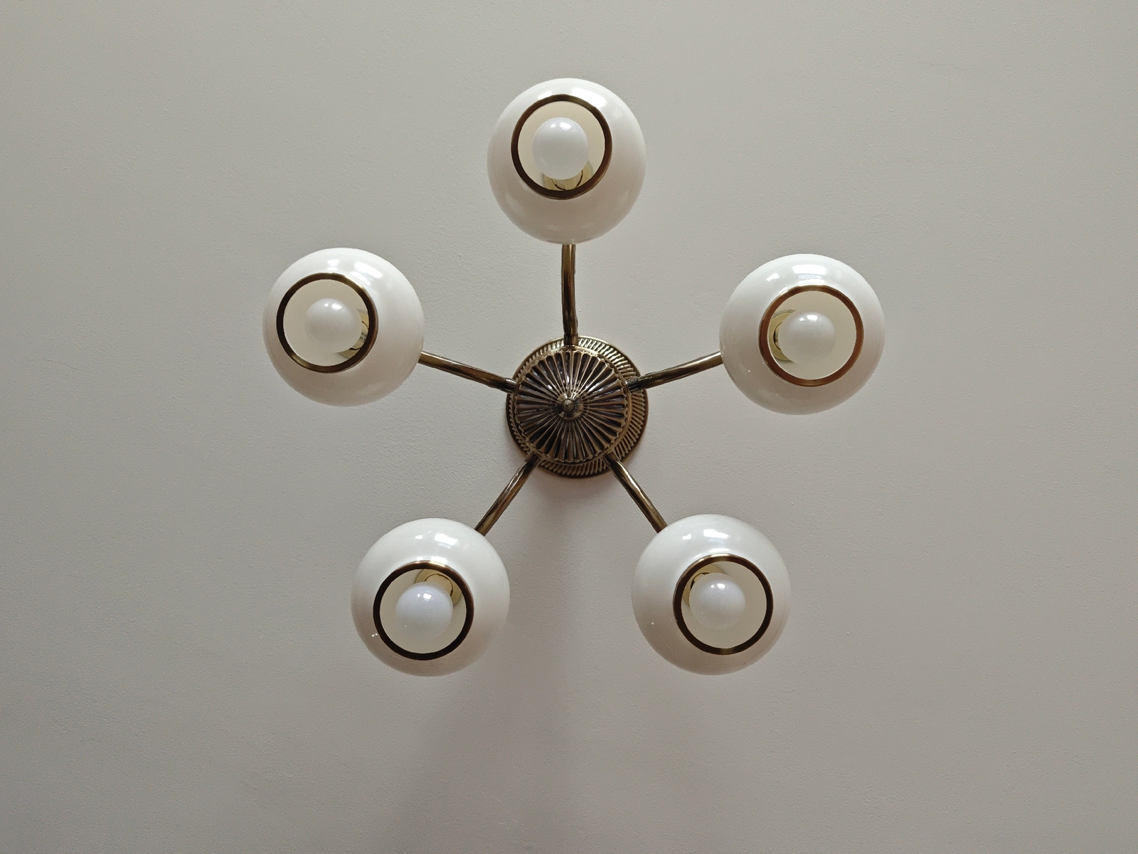 A ceiling-mounted, star-shaped chandelier hangs from a textured, off-white ceiling. The chandelier has a brass-colored central fixture with intricate detailing. Five arms extend from the central fixture, each supporting a white, spherical globe. Inside each globe, a clear glass bulb is visible, illuminated by an unseen light source. The overall aesthetic suggests a vintage or retro design. The image was captured indoors, likely in a residential or commercial space within the city of Tbilisi, Georgia.