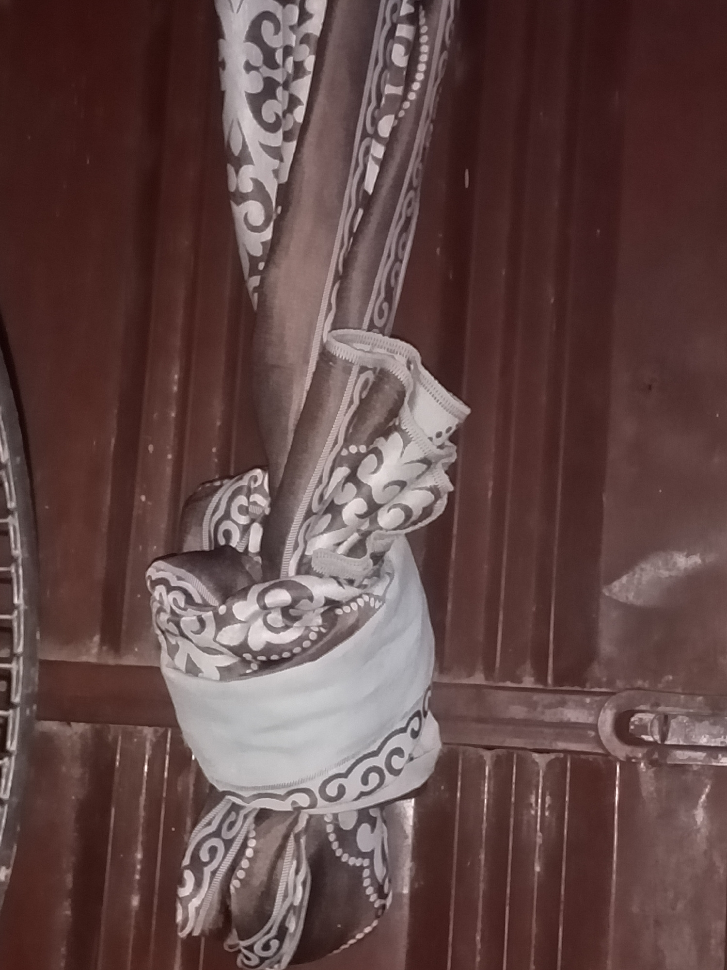 A close-up, vertically oriented, low-angle, slightly blurry photograph shows a piece of fabric tied into a knot against a dark brown, corrugated metallic surface. The fabric is patterned with a repeating white swirl design on a dark brown background, with a white band near the knot. The metallic surface has vertical ridges and appears to be part of a door or gate. The background suggests an outdoor setting, possibly in Jalingo, Nigeria, given the common use of corrugated metal for security and enclosures in the region. There are no people or animals visible in the image. The lighting is dim, casting shadows and highlighting the texture of the fabric and the metallic surface.