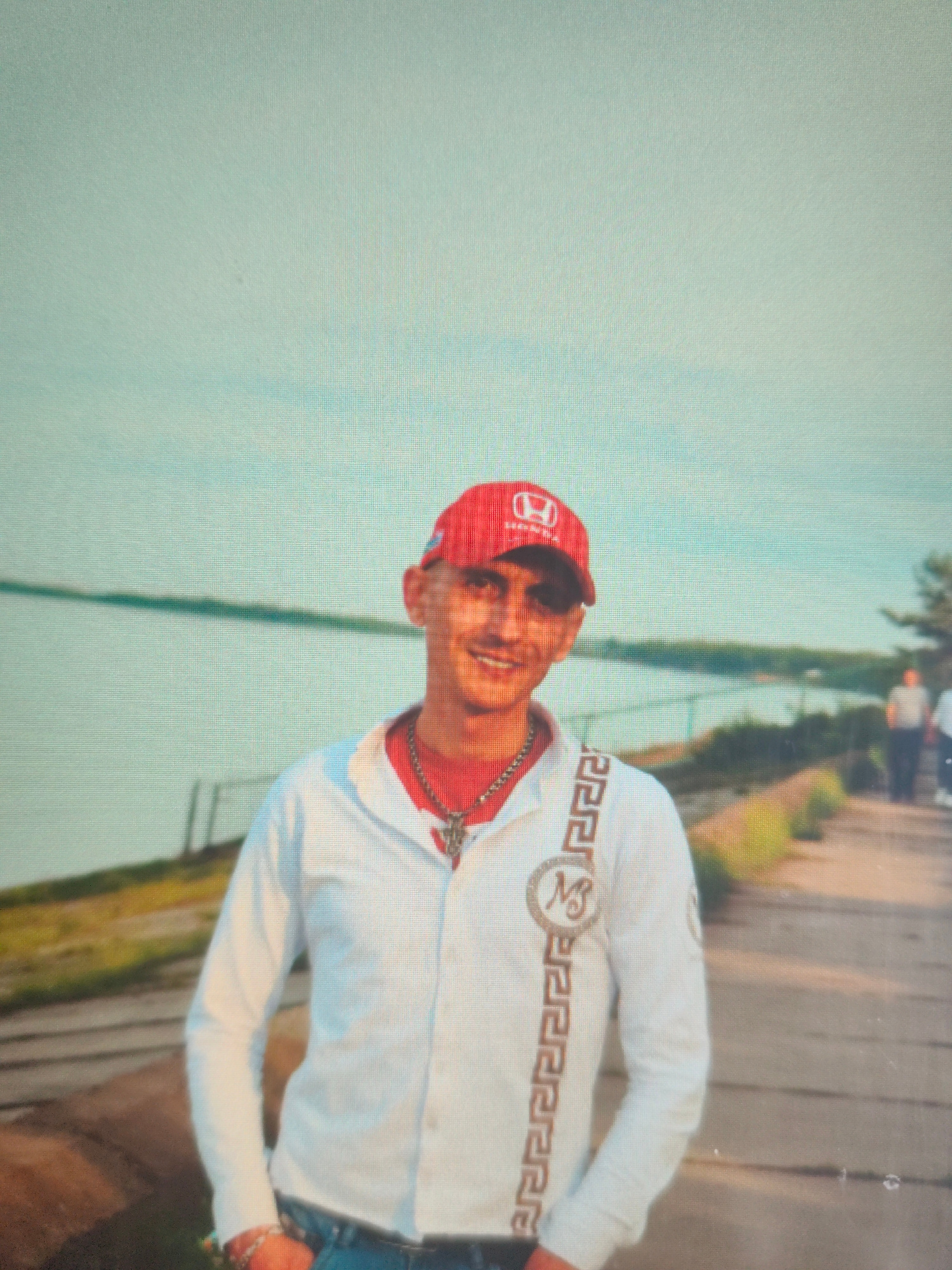A man wearing a red Honda baseball cap, a white collared shirt with a gold emblem and trim, and blue jeans stands in front of a body of water. He is wearing a silver chain with a pendant and a bracelet. The background shows a path next to the water with some vegetation and trees. There are two other people visible in the distance, walking away from the camera. The lighting suggests it is daytime. The image appears to be taken outdoors, possibly in a park or recreational area. The location is identified as the city of Bor, Russia.