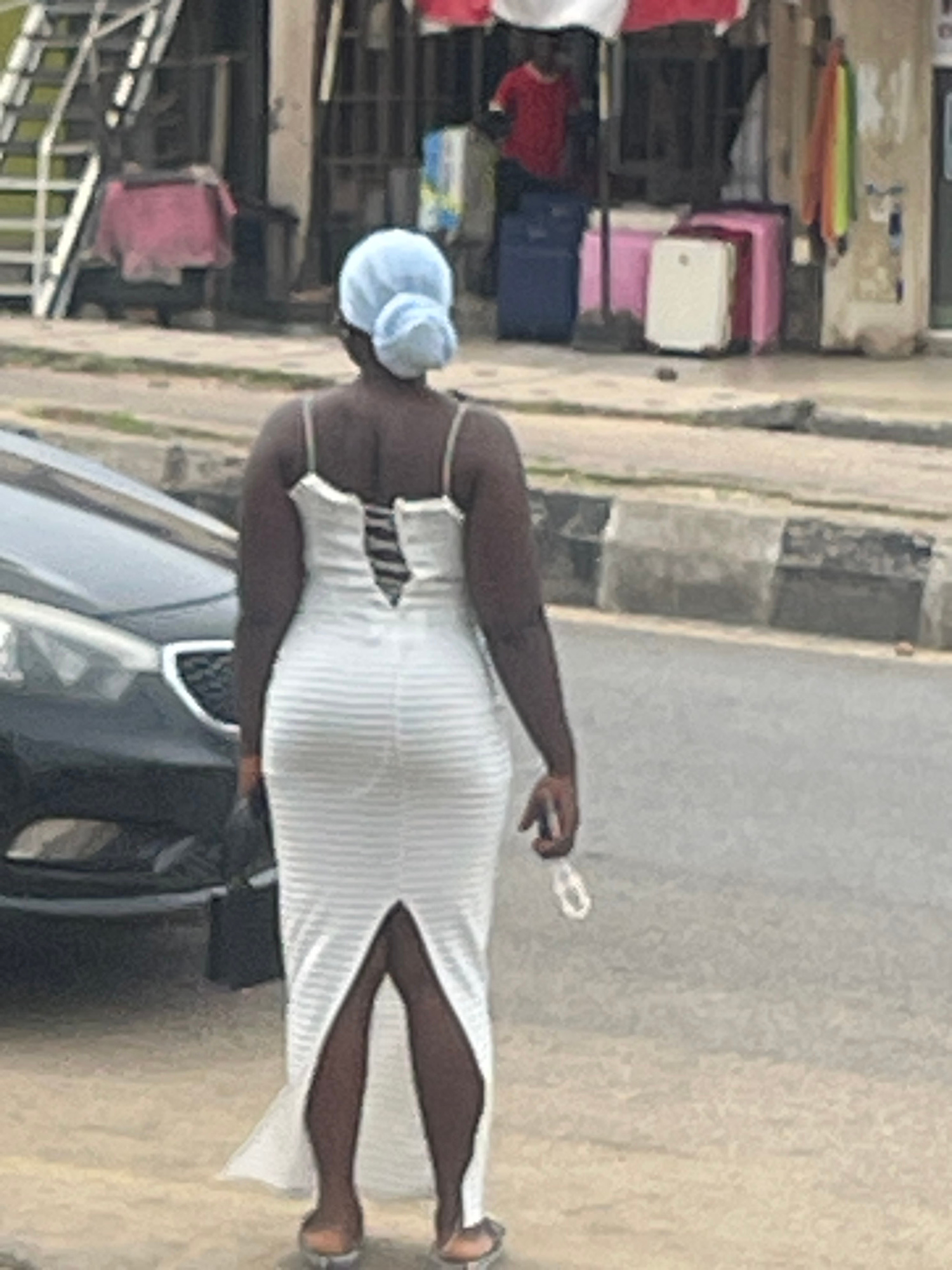 The image shows a person from the back, wearing a white striped dress and a light blue head covering. The person is standing near a vehicle and appears to be holding a small white object in their right hand. The background suggests an outdoor setting, possibly a street with shops or stalls. Colorful items, including what look like stacked suitcases or boxes in pink, red, and blue, are visible on the left side of the image. The lighting suggests it is daytime, but the weather is not discernible. The scene is captured in a slightly blurry, low-resolution manner. There is no visible text in the image.