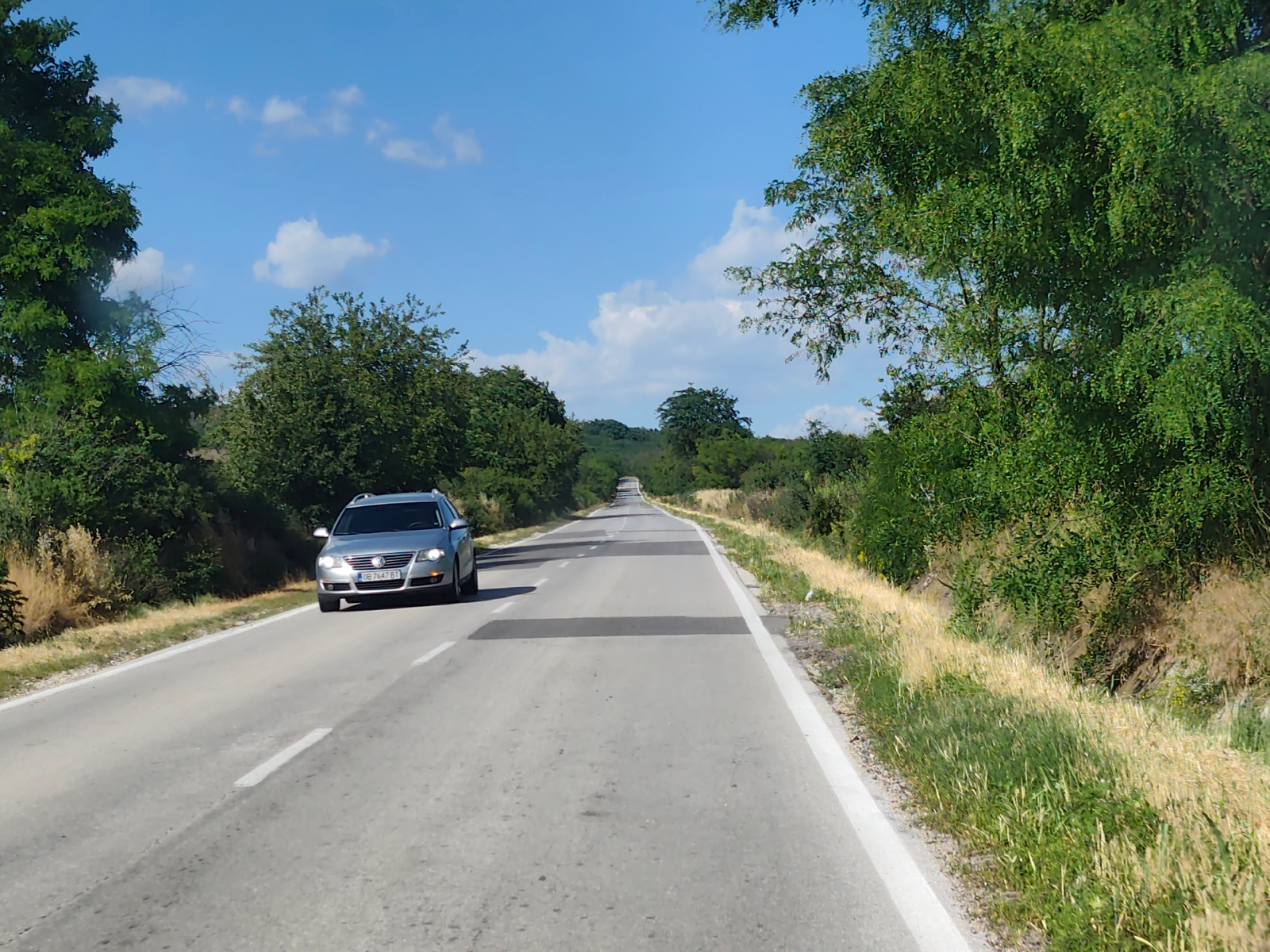 An eye-level, forward-facing perspective captures a silver Volkswagen Passat driving on an asphalt road in Stejerovo, Bulgaria. The car, with license plate OB 7647 BT, is traveling away from the viewer. The road is a two-lane highway with a white solid line on the right edge and a dashed white line on the left, indicating it is a two-way street. The car is in the right lane. The surrounding environment consists of lush green trees and dry grass lining both sides of the road. The sky is blue with scattered white clouds. The scene appears to be during daytime, under clear weather conditions. There are no people visible in the frame besides the implied driver of the car. The road stretches into the distance, curving slightly upwards. Darker rectangular patches are visible on the road surface, possibly for traffic calming or repairs.