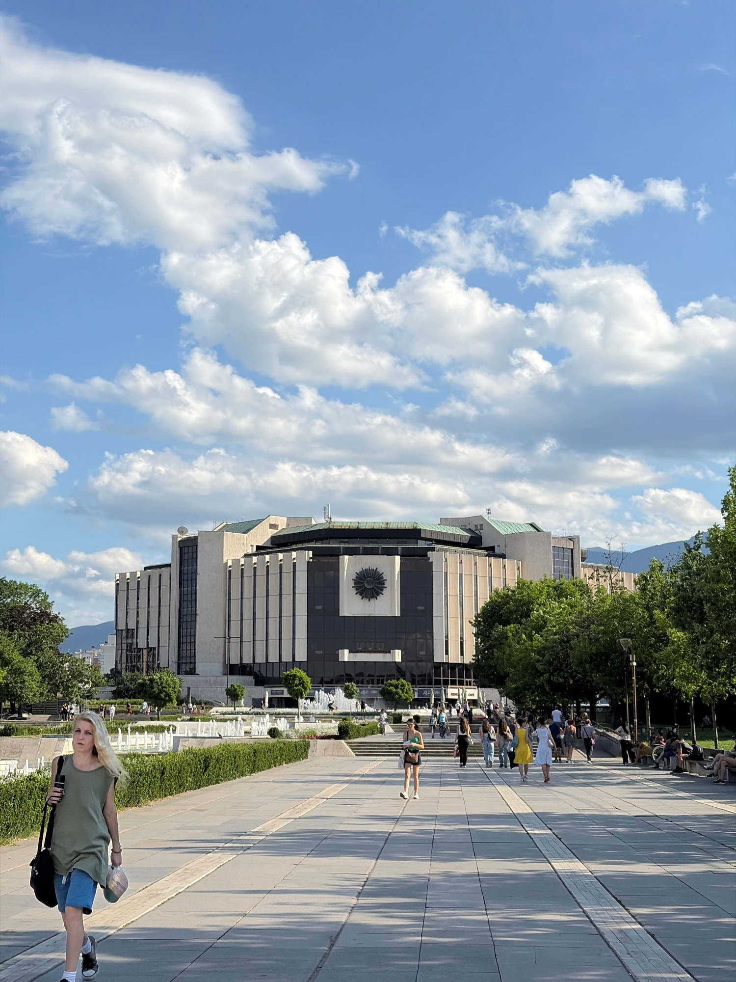 A frontal, eye-level, medium shot captures a sunny day in Sofia, Bulgaria, featuring the National Palace of Culture (NDK) in the background. The NDK is a large, Brutalist-style building with a distinctive circular clock face on its facade. In the foreground, a blonde woman walks towards the left side of the frame, carrying a black bag over her left shoulder and a beige hat in her right hand. She is wearing a sleeveless olive-green top and denim shorts. Other people are visible in the midground, walking on a paved walkway. A row of white fountains stretches across the midground, with lush green bushes lining the walkway. Trees are present on the left and right sides of the frame, and mountains are faintly visible in the distance behind the NDK. The sky is blue with scattered white clouds.