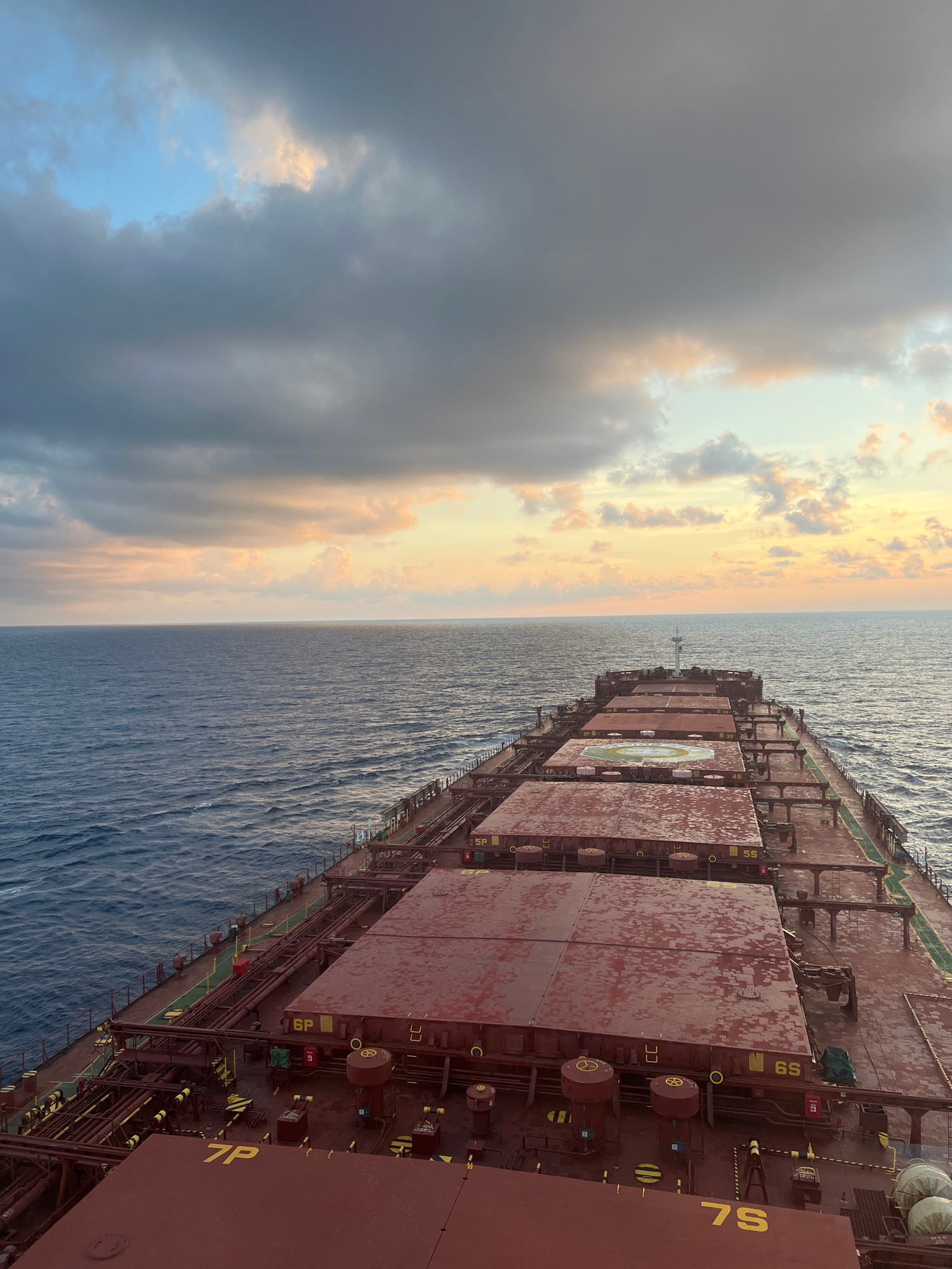 An elevated, forward-looking shot captures the deck of a large cargo ship traversing a vast, calm ocean under a dramatic sky. The sea, a deep, textured blue, stretches to a horizon where it meets a sky painted with a gradient of dusky blues, grays, and soft oranges and yellows, suggesting either sunrise or sunset. Dark, heavy clouds are interspersed with patches of lighter, warmer hues.
The ship's deck is dominated by a series of large, red cargo hatches, some showing the texture of water droplets. Yellow markings and numbers, such as "7P", "6P", "5S", and "6S", are visible on these hatches, indicating specific compartments. Numerous pipes, railings, and metallic structures, all in shades of rust and dark metal, crisscross the deck, leading towards the bow. A helipad, marked with white lines and a circle, is clearly visible on one of the central hatches. The ship appears to be in motion, with a slight wake visible on the port side. No people are visible in the frame. The scene conveys a sense of immense scale and the isolation of being at sea.
