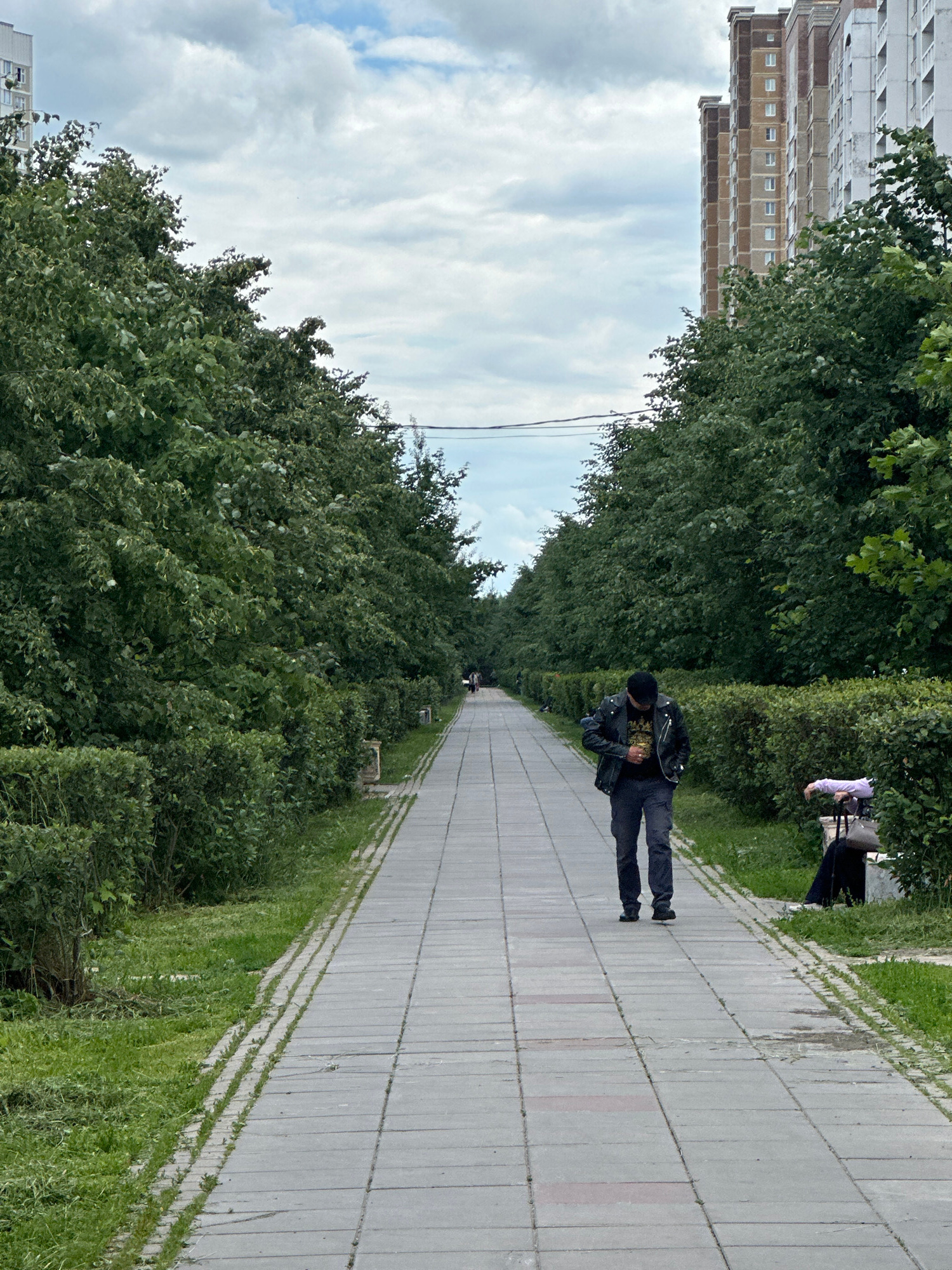 A man in a black leather jacket and black pants walks down a paved path lined with green trees and grass. He is wearing a black beanie and appears to be looking down at something in his hands. In the background, another person in a light-colored shirt is visible, as well as a tall, multi-story building with a beige facade. The sky is cloudy. The scene suggests a daytime walk in a park or urban green space. The perspective of the image is from a tilted angle.