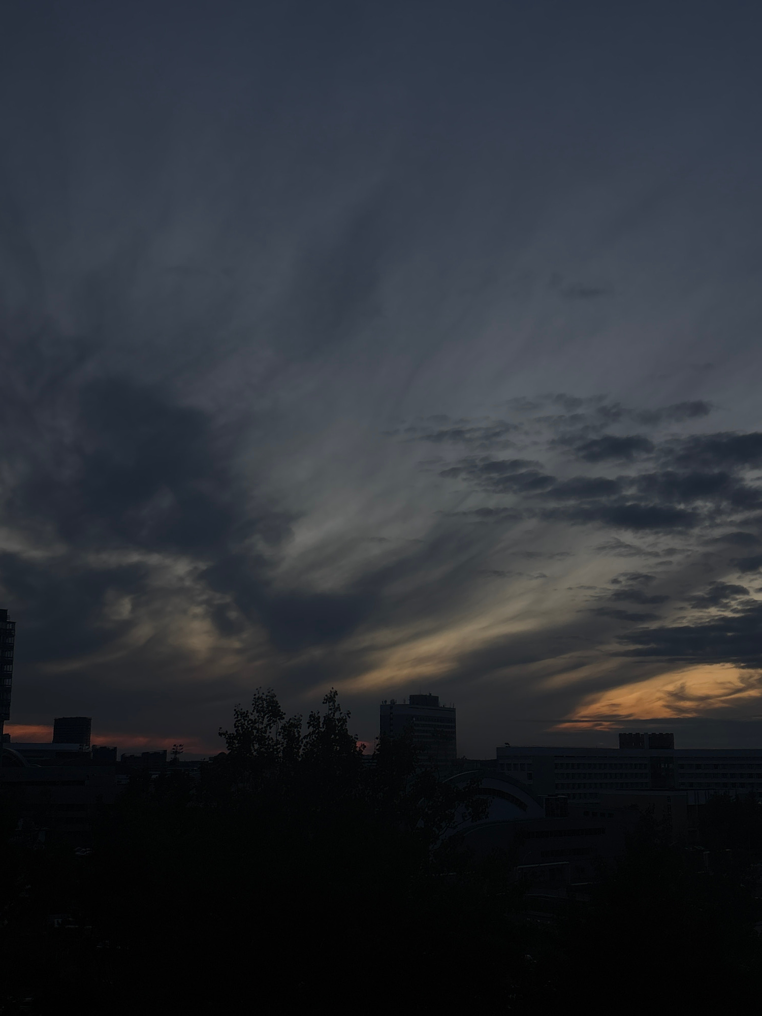 The image displays a dramatic urban landscape at twilight, set against a dynamic and colorful sky. The photograph, taken in Minsk, Belarus, captures a scene primarily dominated by the sky, which fills the upper two-thirds of the frame.
The sky exhibits a compelling contrast of light and shadow. On the left side, the clouds are a dark, deep grey, almost black, indicating the encroaching night. As the gaze moves towards the center and right, the cloud formations become more varied. Darker, more textured clouds are interspersed with lighter, elongated, and wispy streaks that extend diagonally across the sky. These lighter clouds catch the last rays of the low-lying sun, glowing with warm hues of orange, yellow, and pale gold, especially prominent towards the upper right and along the distant horizon.
Below the vibrant sky, on the right side of the frame, several multi-story buildings stand in dark silhouette. Their angular forms create a stark contrast against the illuminated clouds, with faint structural details and windows occasionally discernible where they are touched by ambient light. Dark, dense tree foliage is also visible, intermingled with the buildings, further contributing to the silhouetted urban foreground.
The scene is devoid of people or any specific activity, conveying a sense of stillness and the quiet grandeur of the urban environment transitioning from day to night. The overall mood is atmospheric and contemplative, emphasizing the natural beauty of the sunset or sunrise over a city. There is no visible text in the image.