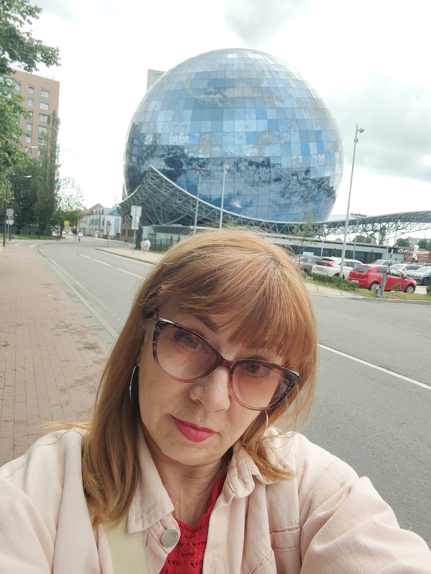 A woman with auburn hair and glasses takes a selfie on a sidewalk. In the background, a large, spherical building with a blue, glass-paneled exterior is visible, reflecting the sky. The building appears to be the Curonian Spit National Park Visitor Center in Kaliningrad, Russia. A street with cars and a sidewalk runs in front of the building. Trees and a multi-story building are also present in the background. The sky is overcast.