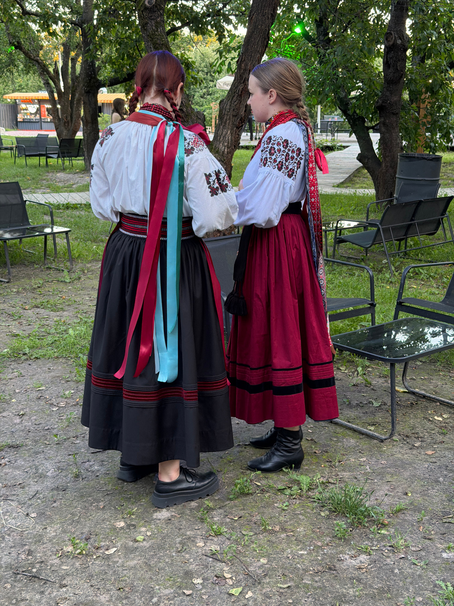 Two individuals, viewed from behind and the side, stand facing each other in a park-like setting. Both are wearing traditional Ukrainian folk costumes. The person on the left has a white embroidered blouse with prominent red and teal ribbons cascading from their neck. They wear a black skirt with red horizontal stripes near the hem, and black leather shoes. The person on the right wears a similar white embroidered blouse with red embroidery and a red scarf around their neck. Their skirt is a deep red with black horizontal stripes near the hem, and they are wearing black boots. They appear to be in conversation or observing something together.
The background consists of trees, grass, and paved ground. Scattered throughout the area are outdoor seating arrangements, including dark metal chairs with dark upholstery, and small dark metal tables with glass tops. In the distance, there is a glimpse of a building with an orange awning and some signs, suggesting a cafe or event space. The lighting indicates it is daytime, possibly late afternoon, with dappled sunlight filtering through the trees. The presence of picnic tables and chairs implies a public outdoor gathering space, typical of urban parks. This scene is consistent with a location in Kyiv, Ukraine, given the traditional attire and the style of the park amenities.