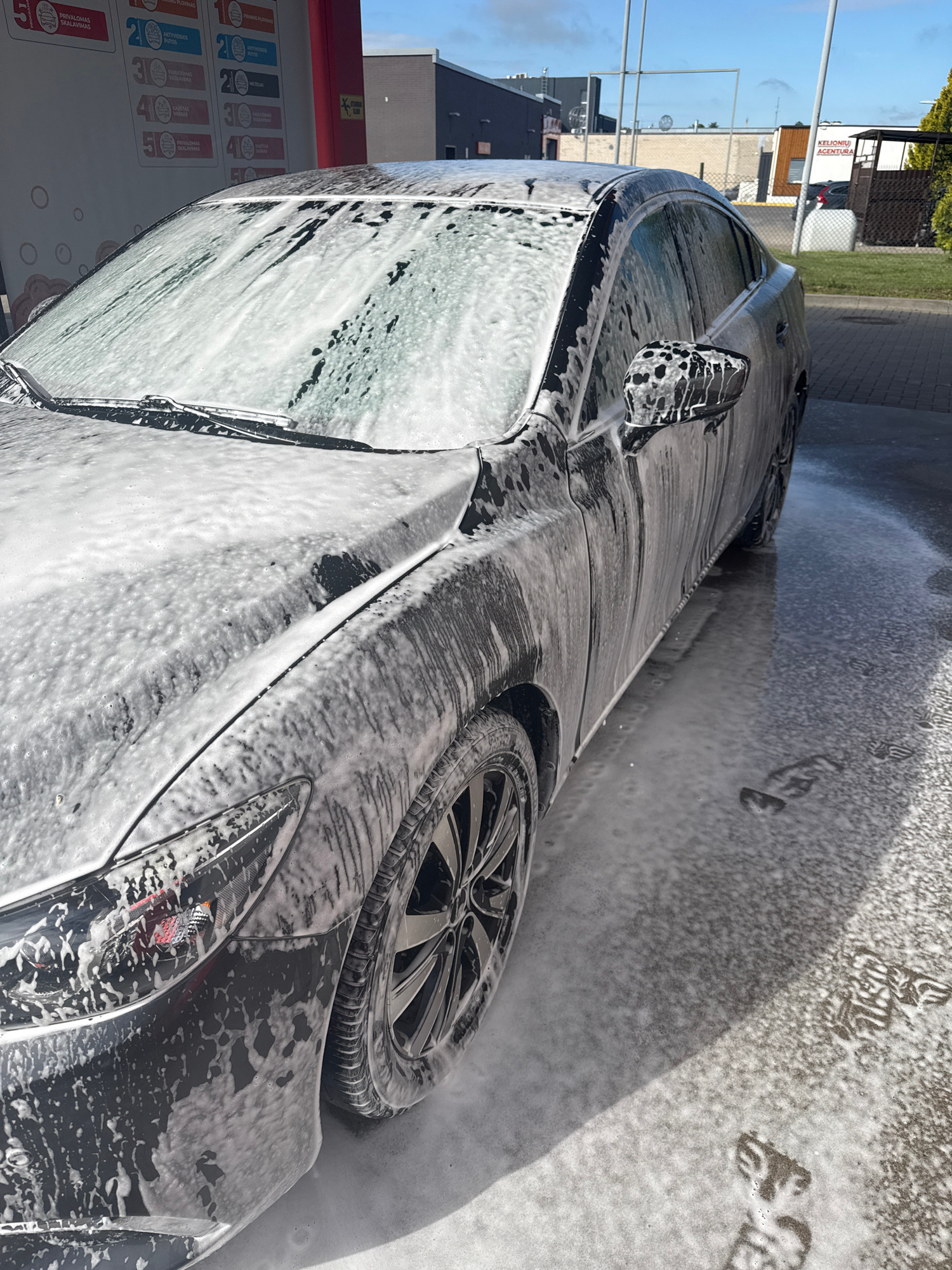 An outdoor, low-angle, eye-level, medium shot captures a dark sedan at a self-service car wash station. The car is covered in a thick layer of white foam, with water streaks running down its sides and windshield. The vehicle is positioned on a paved surface, with puddles of soapy water on the ground. In the background, various buildings and a clear sky are visible. A sign with lettering in an unknown language is attached to a red structure to the left of the car. The scene is well-lit by natural sunlight.