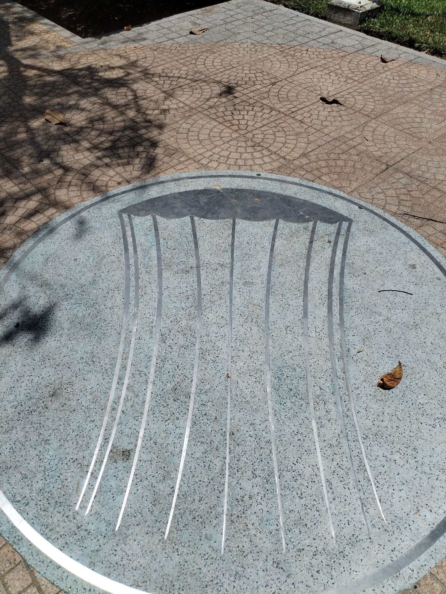 The image shows a circular, light blue, speckled surface with several thin, reflective metal strips embedded in it. These strips are arranged in a radial pattern, converging towards the center of the circle. A shadow of what appears to be tree branches is cast across the surface, indicating an outdoor setting under sunlight. The surface is also speckled with small dark particles, and there are a few fallen brown leaves scattered on it, further suggesting an outdoor environment. The ground surrounding the circular object is paved with interlocking cobblestones in a circular pattern.
To the top and left of the circular surface, the paving is darker and appears to be a pathway, with shadows of tree leaves also cast upon it. To the right, there is a patch of green grass. The overall lighting suggests it is daytime. The scene is tranquil and appears to be in a public outdoor space, possibly a park or plaza in Nha Trang Ward, Vietnam. There is no visible text in the image.