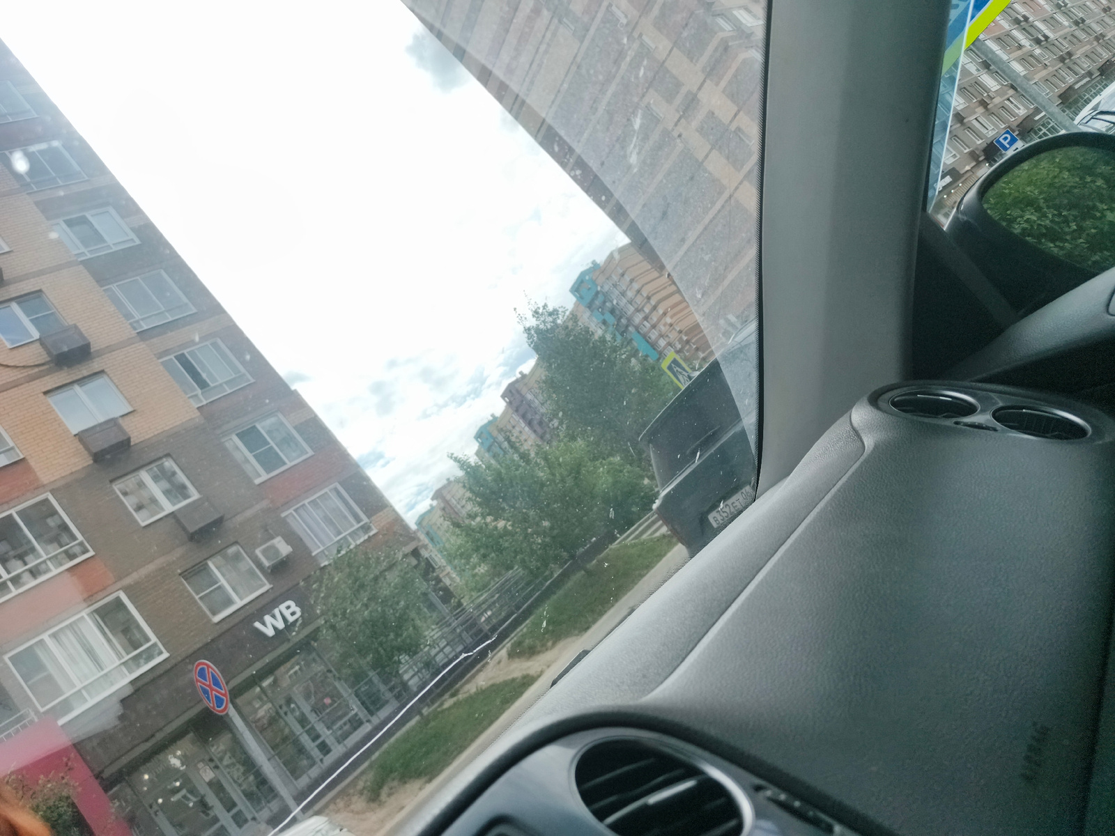 The image shows a view from inside a car, looking out through the windshield. The car is parked or driving on a street in a residential area. On the left, there is a multi-story brick building with many windows. Below the building, a sign with "WB" is visible, along with a circular sign with a red border and a horizontal line. Trees are visible in front of and around the building. Further in the background, more buildings with colorful facades and apartment windows can be seen, along with a cloudy sky. On the right side of the image, part of the car's interior is visible, including the dashboard and a side mirror. A license plate with Russian characters and numbers is visible on a car in the background. The overall scene suggests a daytime setting, likely a residential neighborhood.