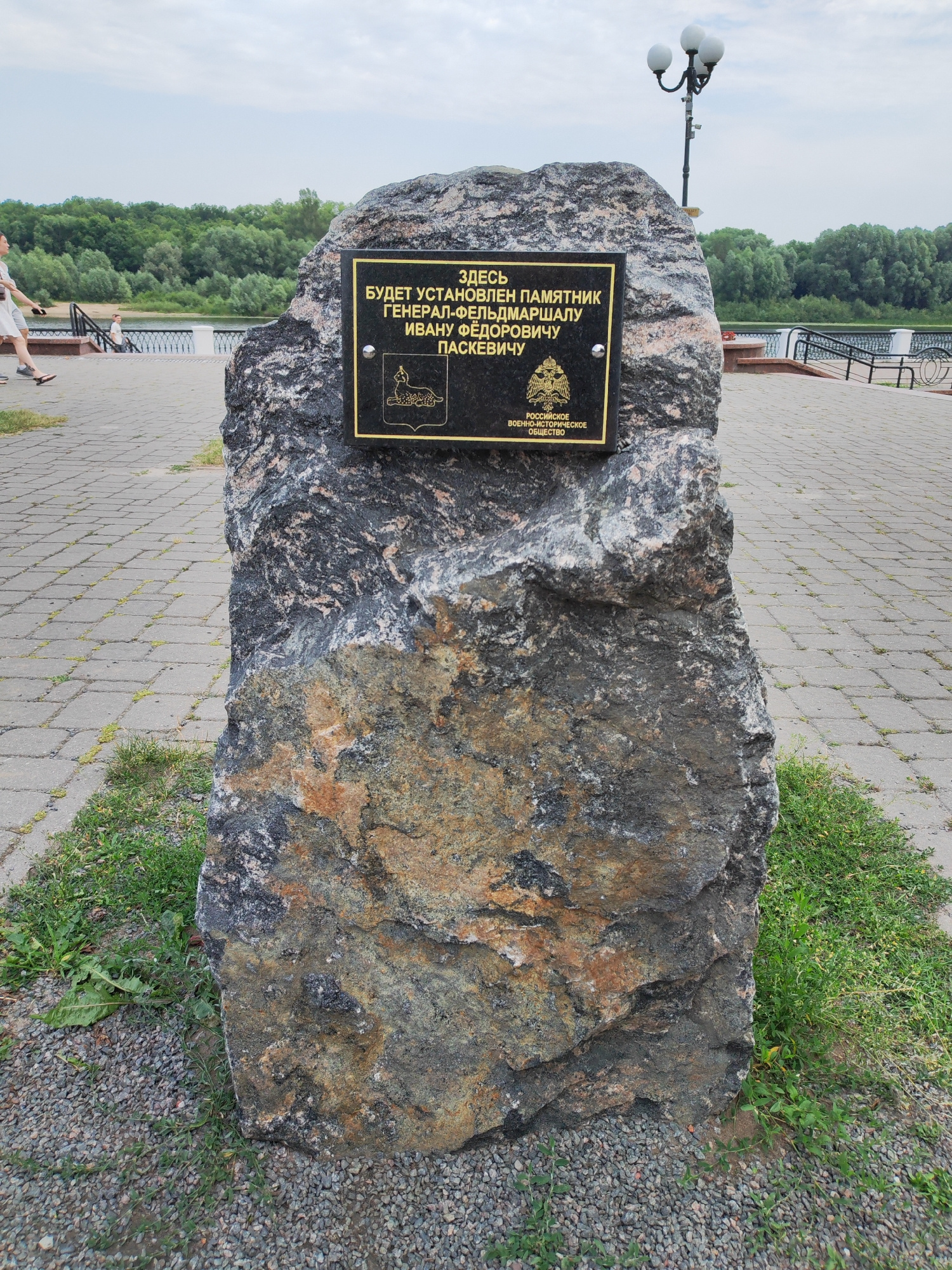 This is a vertical, eye-level, outdoor photograph of a stone monument with a commemorative plaque. The monument, made of rough, dark grey and reddish-brown stone, is the central focus. Attached to its face is a black plaque with gold lettering. The text on the plaque, in Russian, reads "HERE WILL BE ERECTED A MONUMENT TO GENERAL-FIELD MARSHAL IVAN FYODOROVICH PASKEVICH" and also mentions the "RUSSIAN MILITARY-HISTORICAL SOCIETY". Below the main text are two emblems.
Behind the monument, a paved walkway is visible, made of grey interlocking bricks. To the left of the monument, a body of water, likely a river, is seen with a treeline on the far bank. A lamppost with three globes stands to the right of the monument, further back. A few people are walking on the path in the background, one in a light-colored dress and another in darker clothing. The sky is overcast, suggesting a cloudy day. The overall scene appears to be a public park or embankment, likely in Homyel, Belarus, given the text on the monument which refers to a local historical figure. The image captures a moment of anticipation for the erection of the monument.