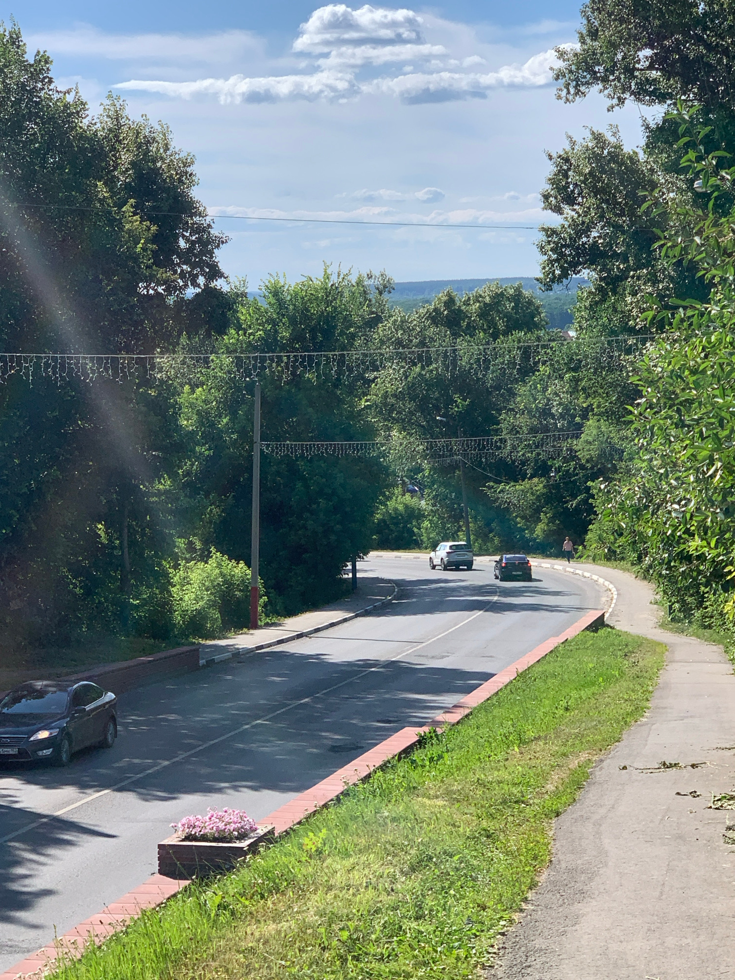 This is a photograph taken from a high vantage point overlooking a curved road lined with trees and grass. Two cars are visible on the road. The sun is shining brightly, casting shadows on the road and grass, suggesting it is daytime. The sky is blue with some white clouds. The presence of trees, grass, and a road suggests an outdoor, possibly rural or suburban setting. There is a flowerbed with pink flowers visible on the side of the road. The image has been rotated 90 degrees counter-clockwise. No people are clearly identifiable.