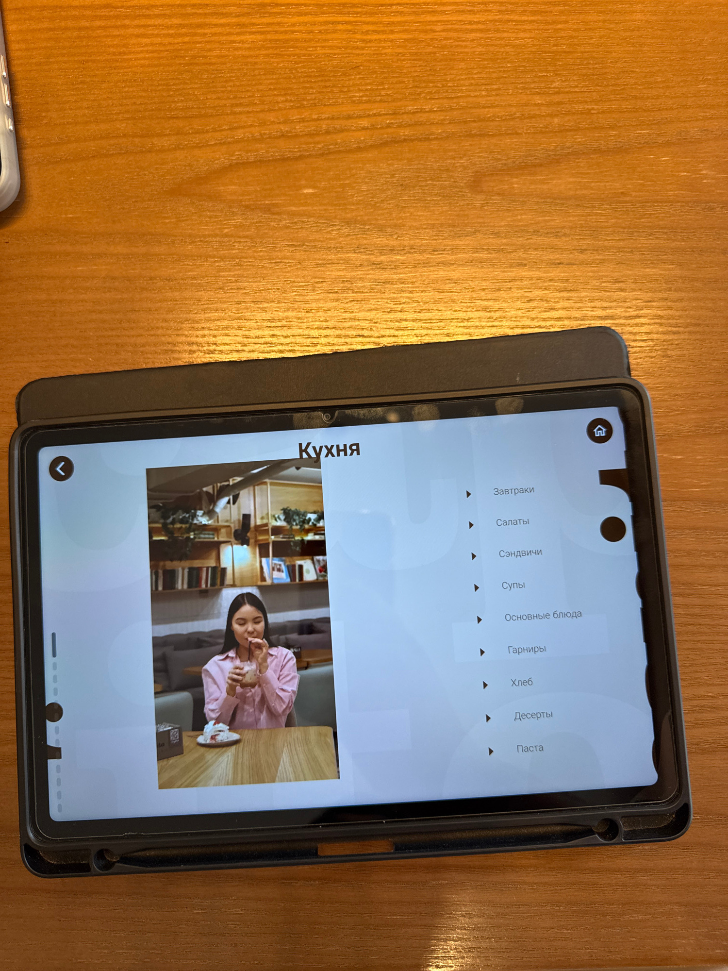 A tablet is placed on a wooden surface, displaying a user interface with menu options. The screen shows the title "Кухня" (Kitchen) at the top. Below the title, a photograph of a young woman in a pink shirt is visible, holding a drink with a straw and sitting at a table with a slice of cake. To the right of the photograph, a list of menu categories is displayed in Russian, including "Завтраки" (Breakfasts), "Салаты" (Salads), "Сэндвичи" (Sandwiches), "Супы" (Soups), "Основные блюда" (Main courses), "Гарниры" (Side dishes), "Хлеб" (Bread), "Десерты" (Desserts), and "Паста" (Pasta). A home icon is present at the top right of the screen, and a back arrow icon is at the top left. The scene does not provide any information or visual cues that would allow for the determination of its location in Almaty, Kazakhstan.
